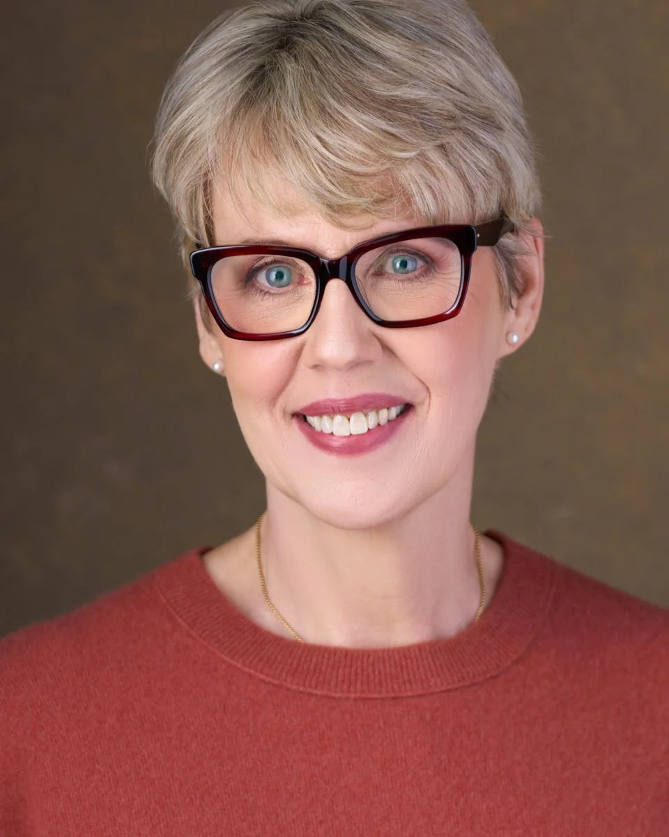 Portrait of a woman with short blond hair, wearing glasses, earrings, and a red sweater, smiling with a brown background.