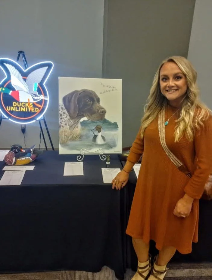 Woman in orange dress standing next to a table displaying a painting of a dog and ducks, with a neon sign reading 'Ducks Unlimited' in the background.