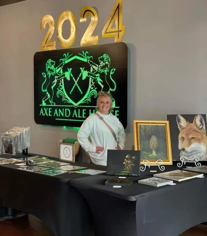 A woman standing behind a table at an art and home decor event in 2024, with various framed artworks displayed, including a fox portrait and a framed mushroom illustration. The background features a large illuminated sign that reads 'AXE AND ALE HOUSE' with a crest of two lions holding crossed axes, flanked by large golden numbers '2024'.