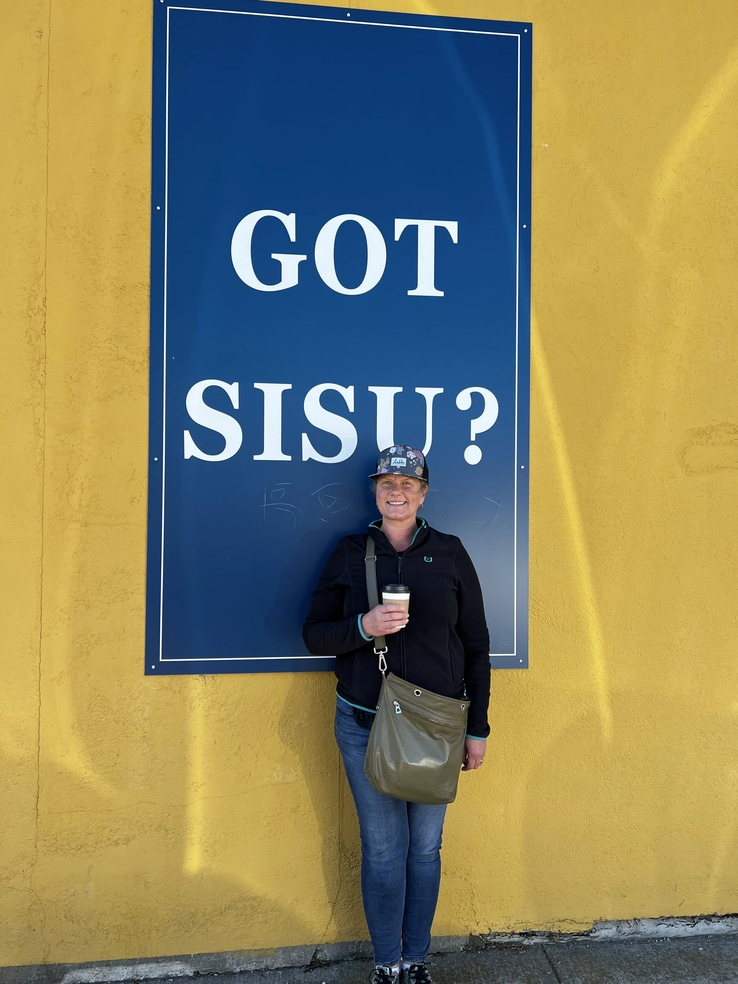 A woman standing in front of a large blue sign with white text that reads 'GOT SISU?' She is smiling, wearing a black jacket, blue jeans, a cap, and holding a coffee cup.