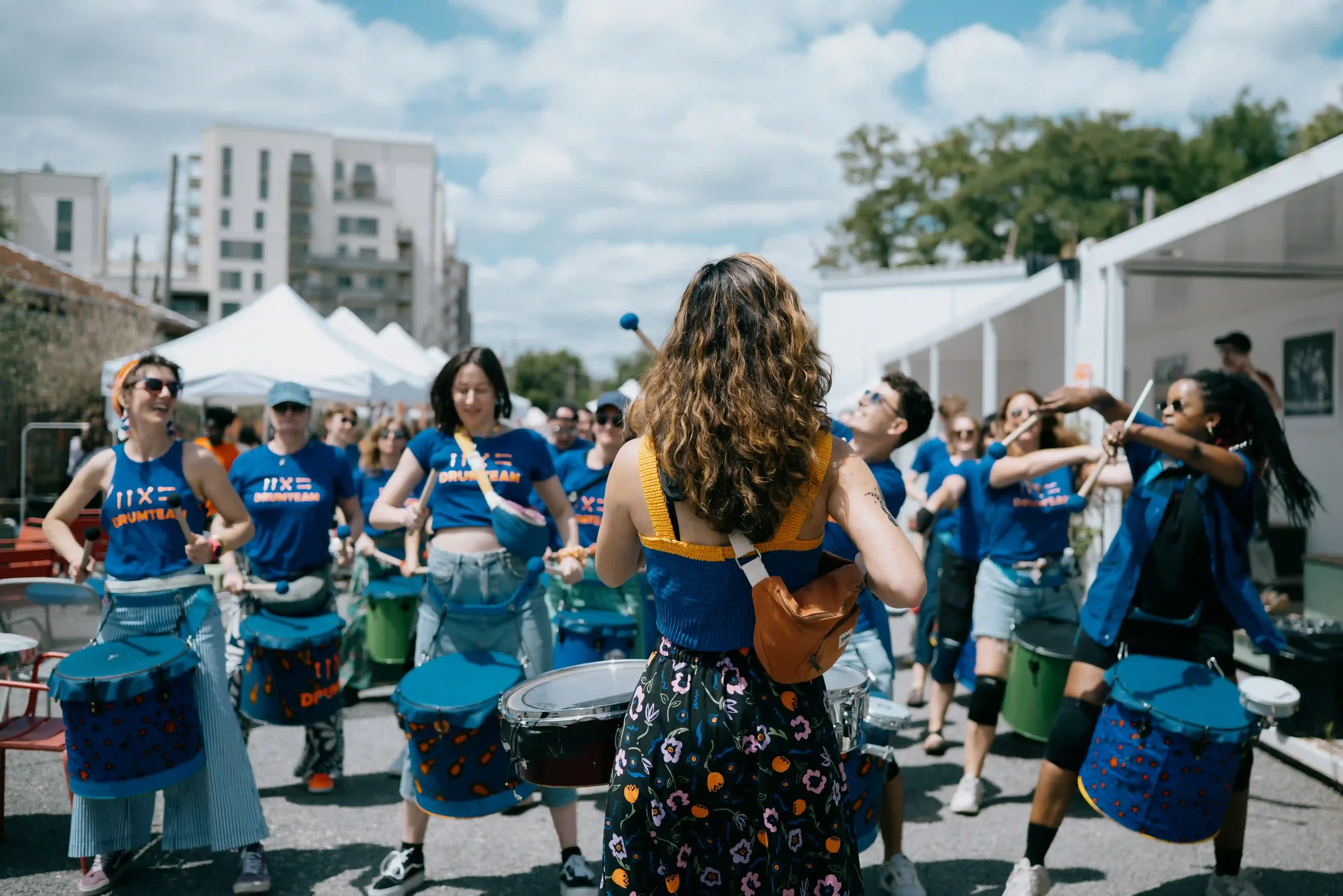 Groupe diversifié de la Drumteam en pleine performance collective lors du festival Emmaus Solidarité, sous un ciel de festival d'été. Photo : Michael Mendes