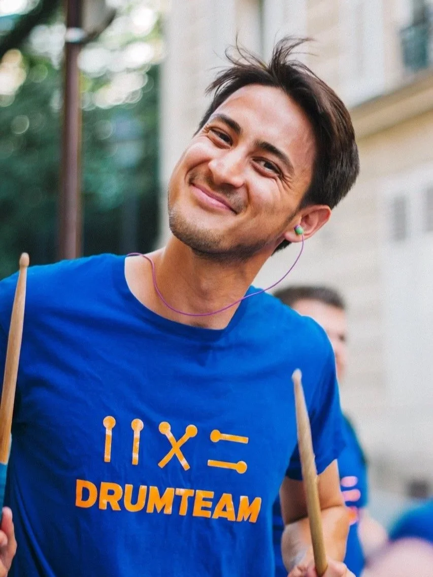 Un homme souriant portant un t-shirt bleu avec le texte 'DRUMTEAM' et jouant de la batterie lors de la Fête de la musique 2025