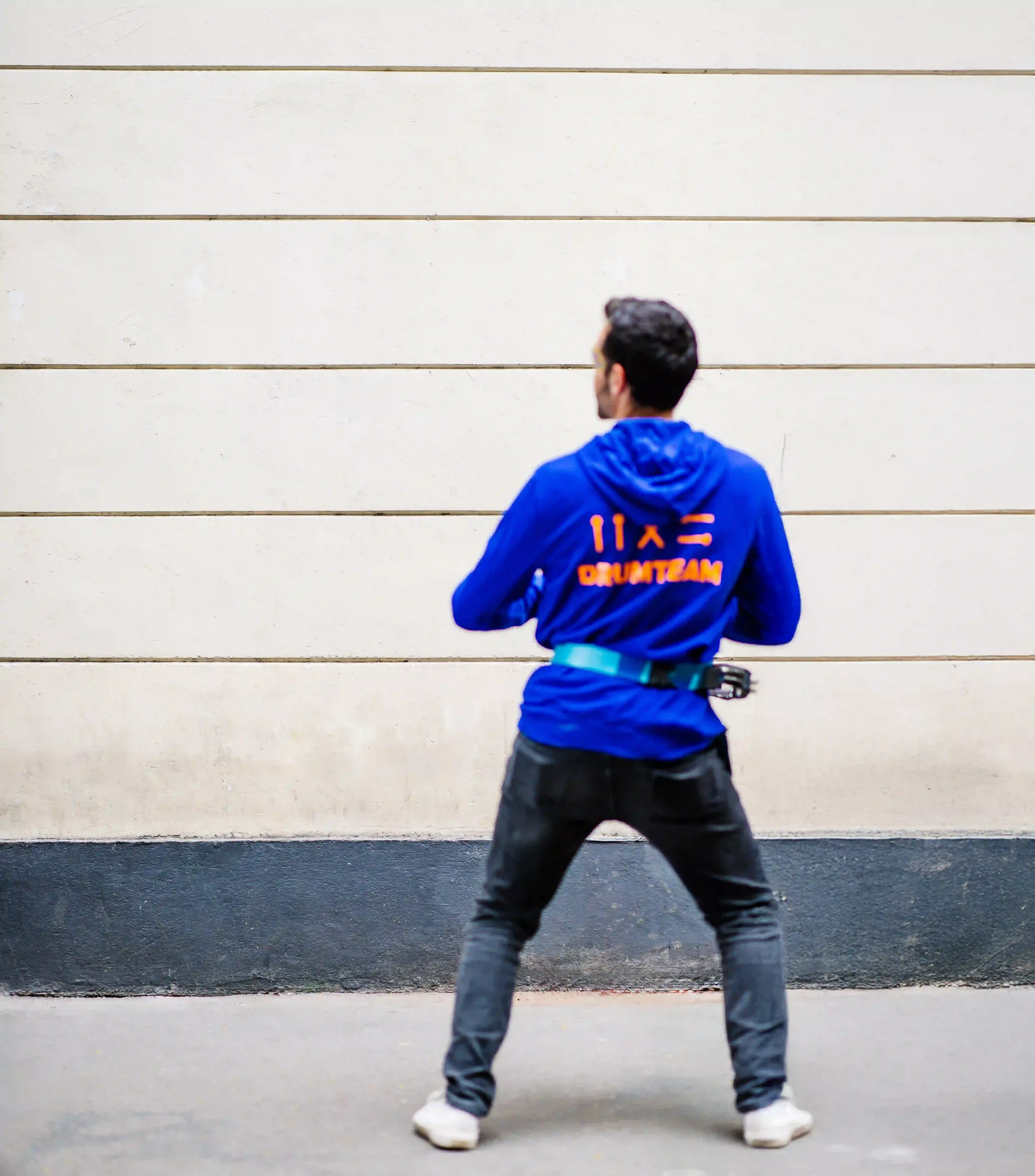 Un homme debout, vu de dos, portant un sweat bleu avec des inscriptions en orange, dans un environnement urbain.