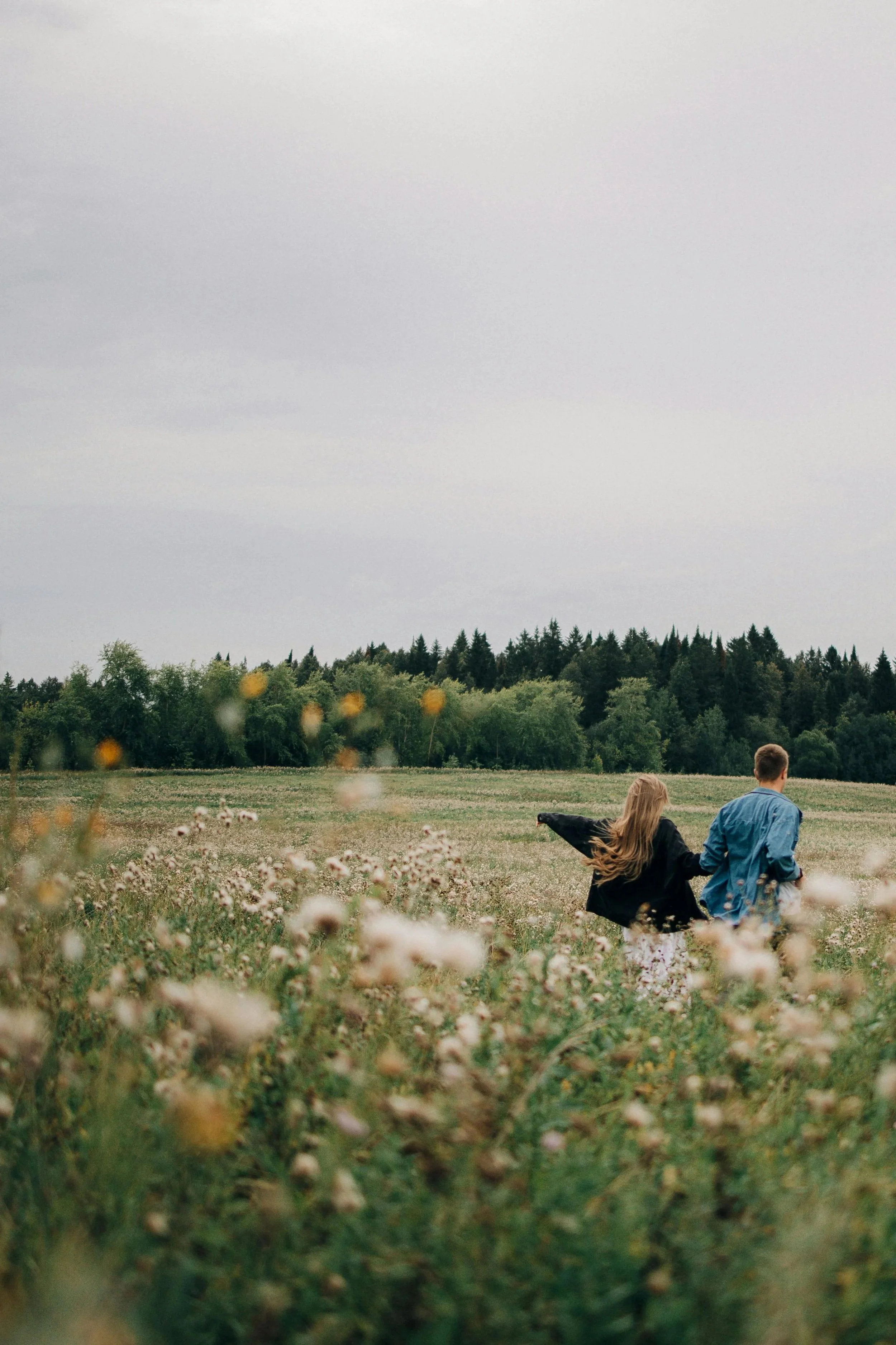 A young man and woman walk through a grassy, flower-filled field under an overcast sky, with a line of trees in the background.