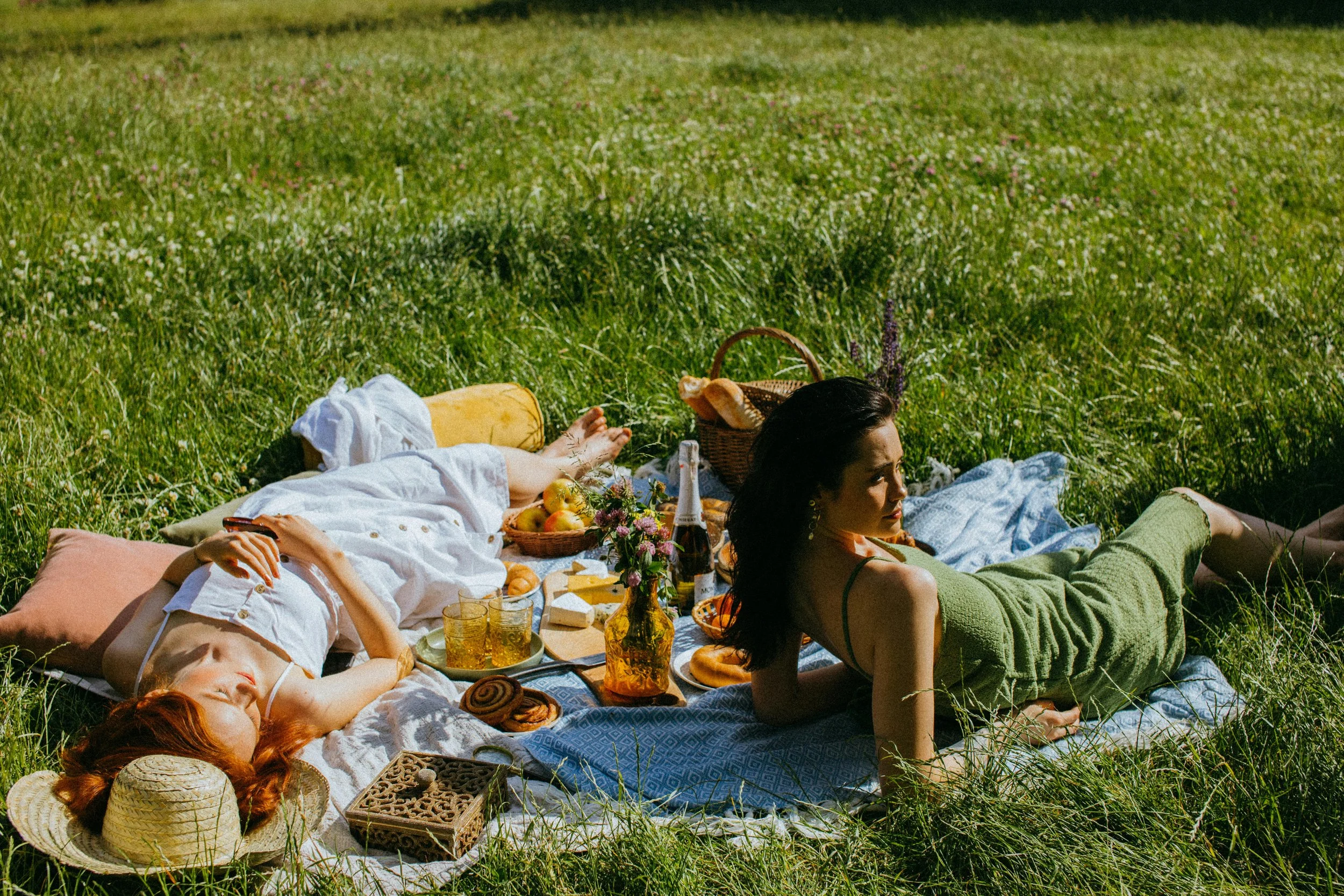 Two women having a picnic on a sunny day in a grassy field. One woman, with dark hair, is lying on a blue blanket, and the other, with red hair, is reclining on a pink pillow. The picnic includes apples, bread, cheese, wine, and snacks in baskets and on plates.
