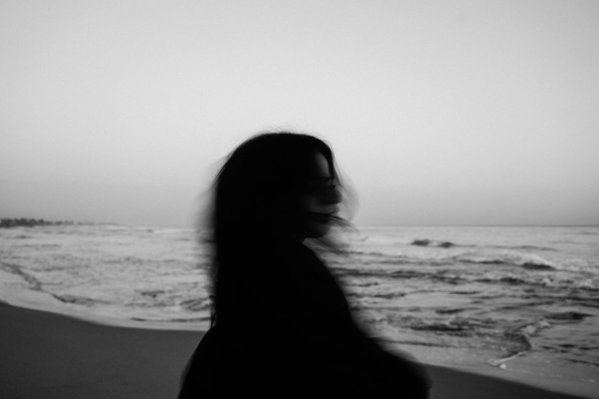 A blurred black and white photo of a woman with long hair standing on a beach, facing the ocean during sunset or sunrise.
