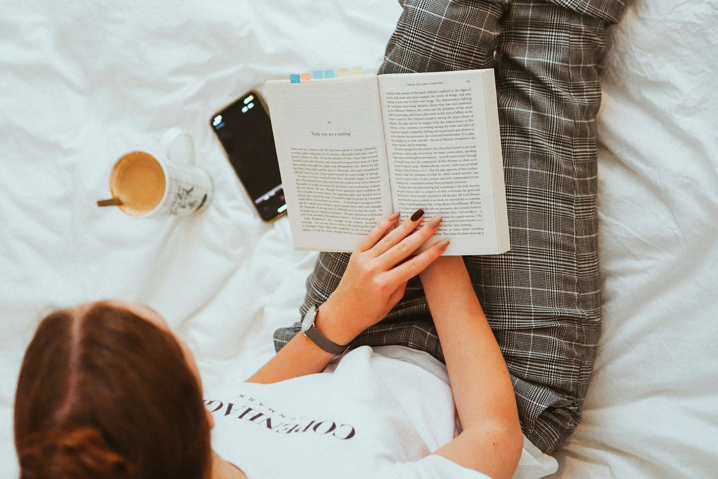 A person wearing plaid pants is sitting on a bed reading a book, with a drink and a phone nearby.