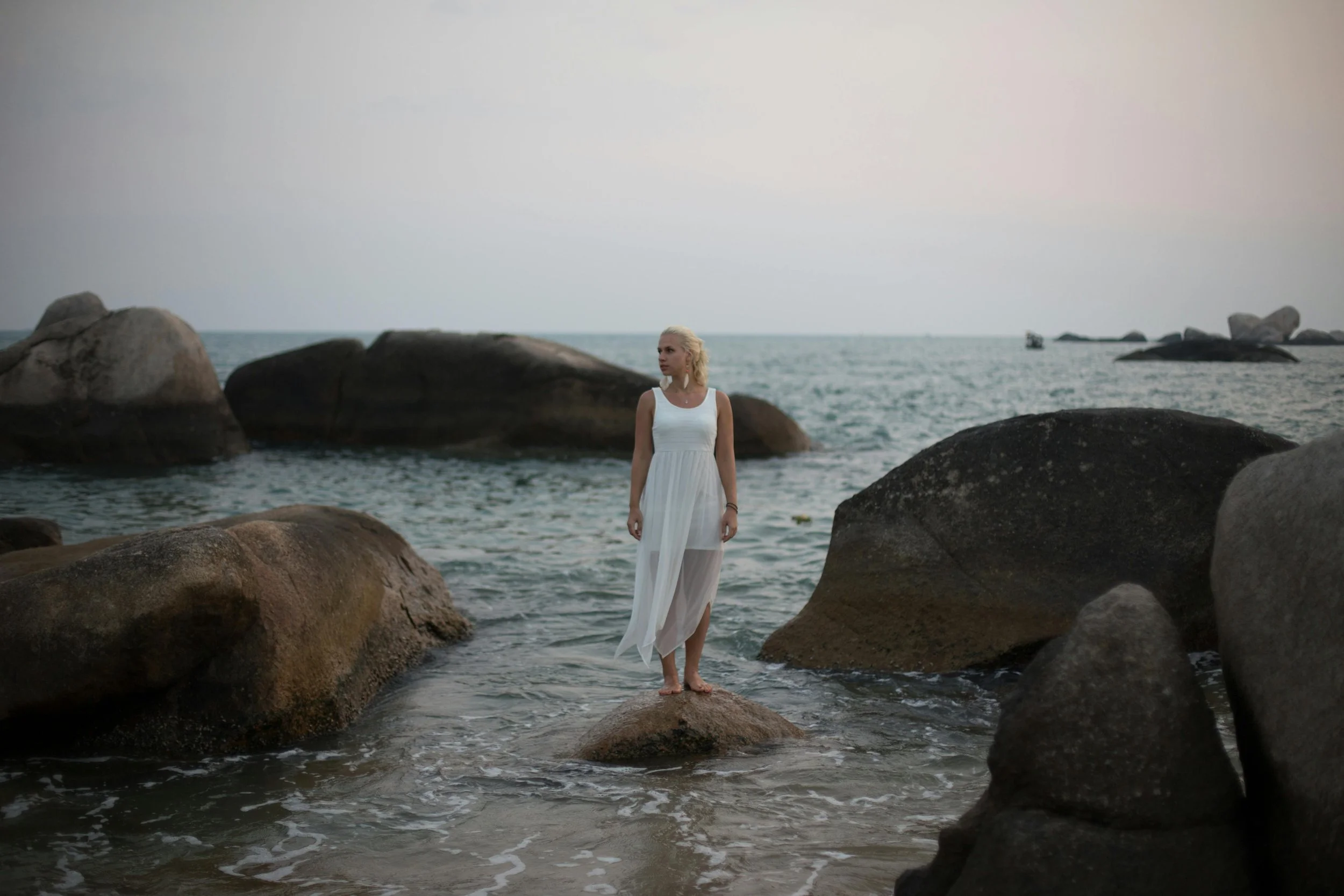  Woman in white dress standing on coastal rocks surrounded by water, looking out to sea
