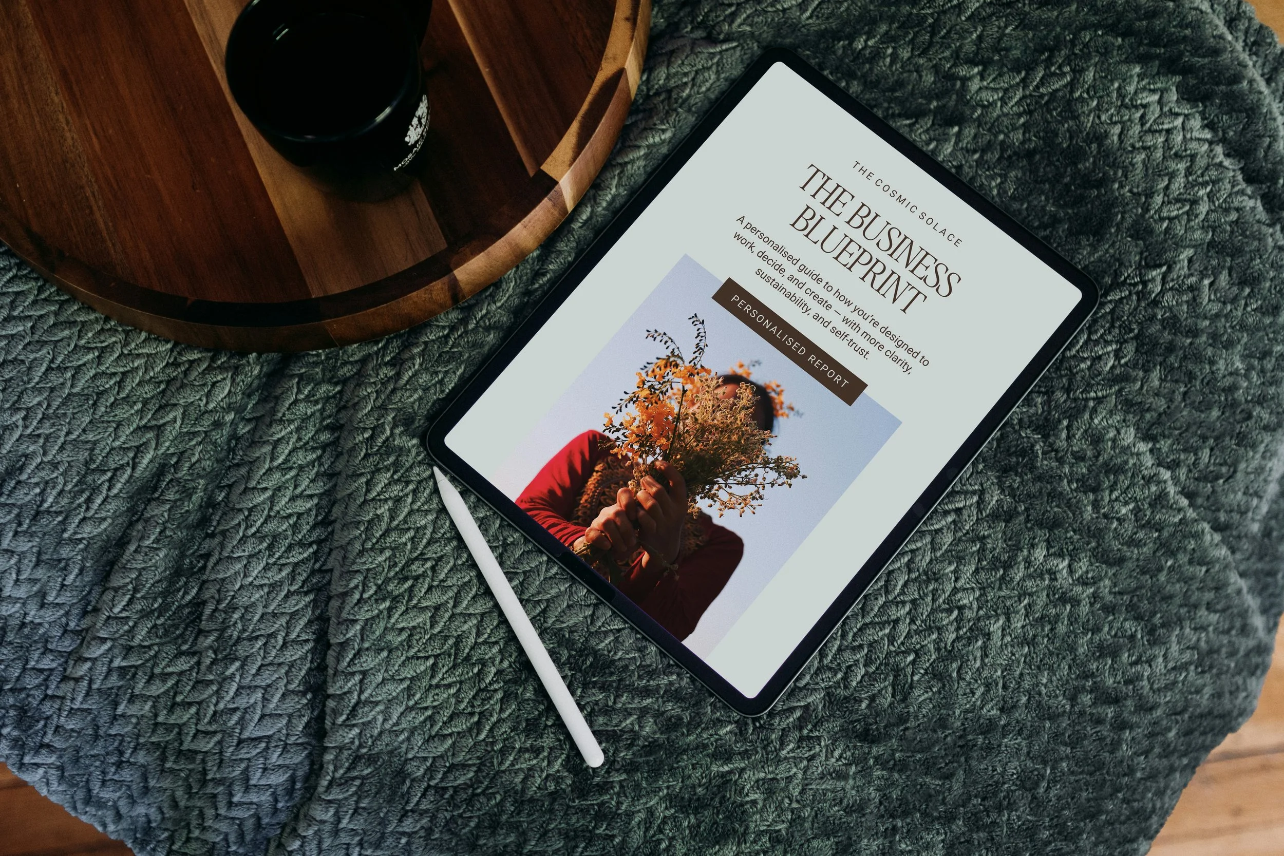 A tablet displaying a digital guide titled 'The Business Blueprint' with a subtitle about designing work for sustainability and self-trust, resting on a textured gray blanket. Nearby, a white stylus pen. To the left, a round wooden tray holds a black cup.