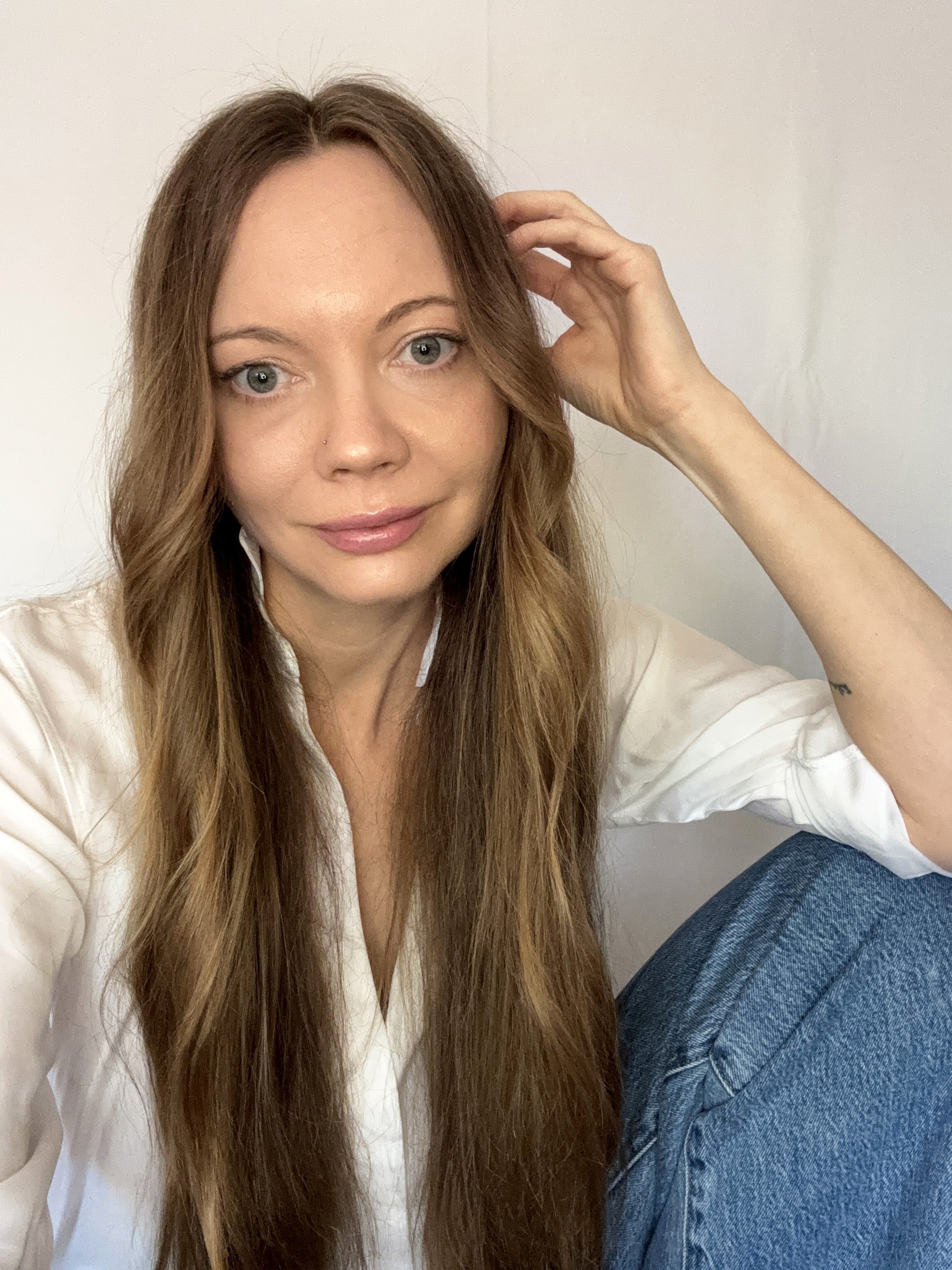A woman with long, wavy brown hair and blue eyes, wearing a white blouse and blue jeans, sitting against a plain white wall.