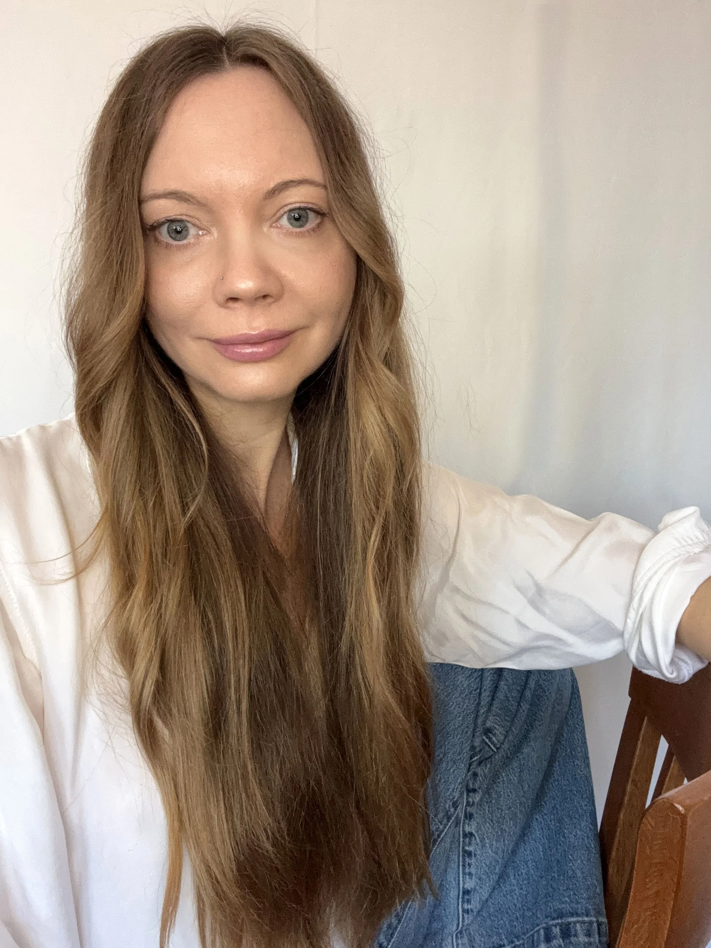 A woman with long, wavy, light brown hair, blue eyes, and light skin, taking a selfie indoors.
