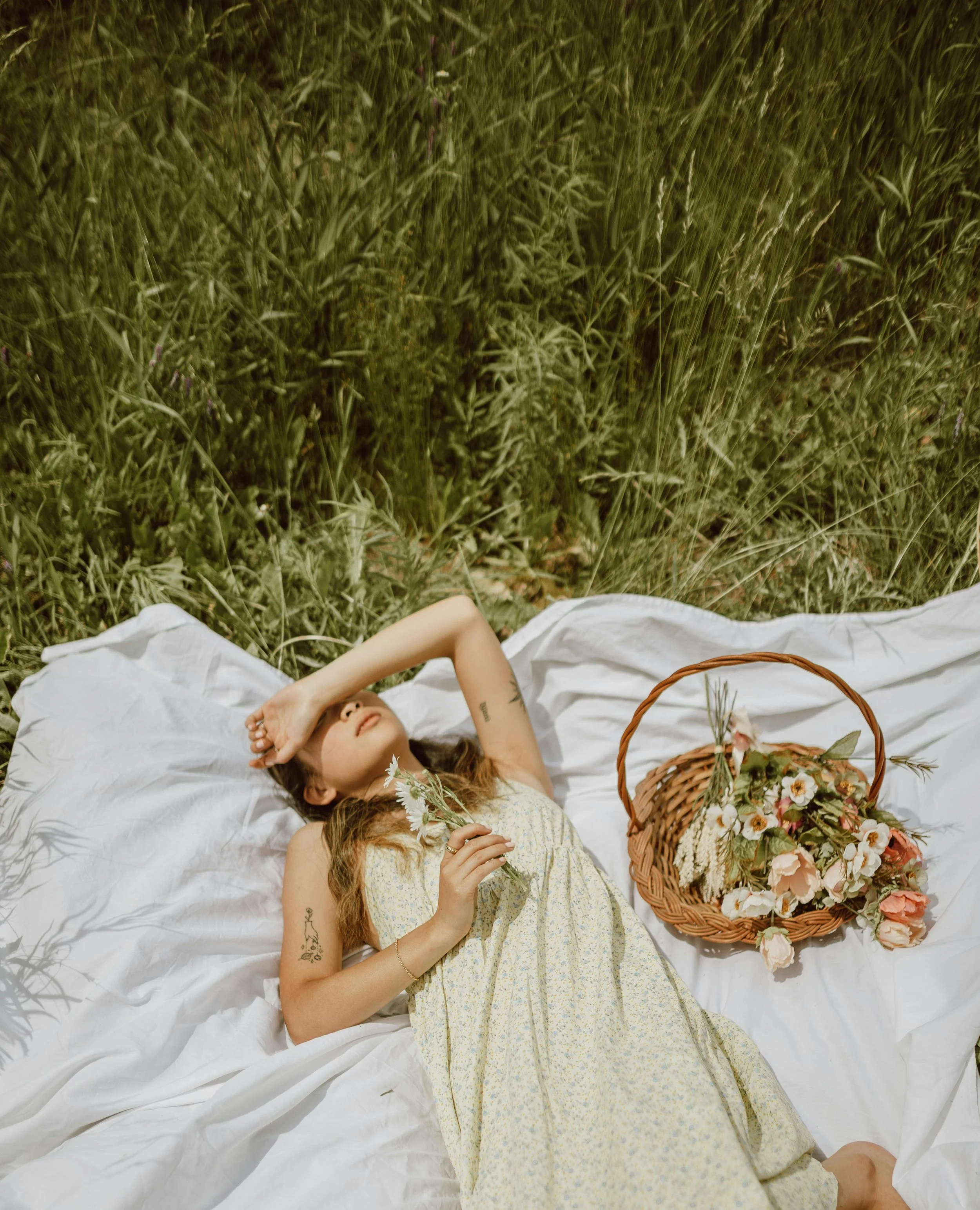 A woman lying on a white blanket in a field of tall grass, holding a small bouquet of wildflowers, with a wicker basket filled with flowers nearby.