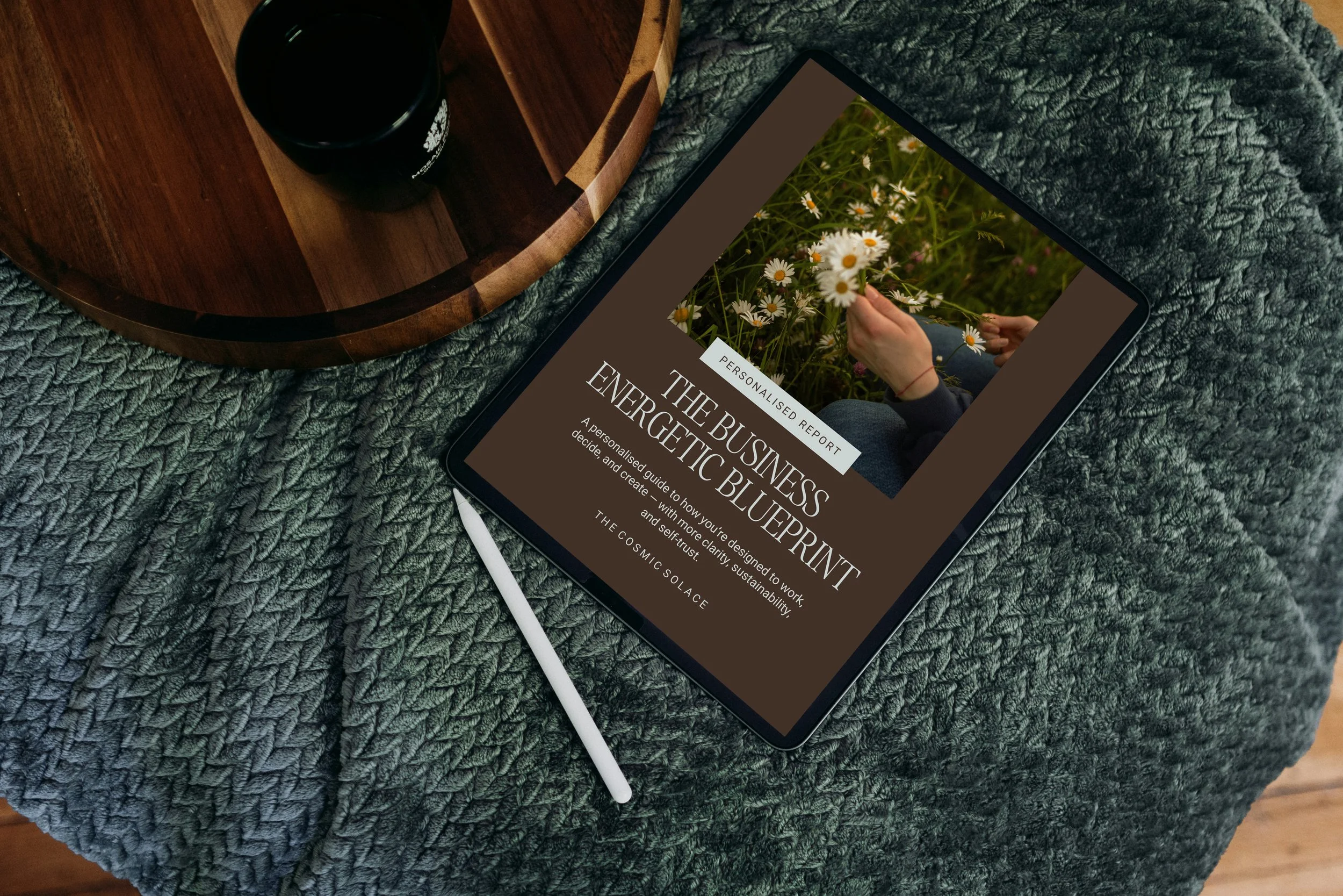 Tablet on a textured green blanket displaying a report titled 'The Business & The Business BluePrint' with a photo of daisies and a person's hand, alongside a white stylus, next to a wooden tray with a dark beverage.