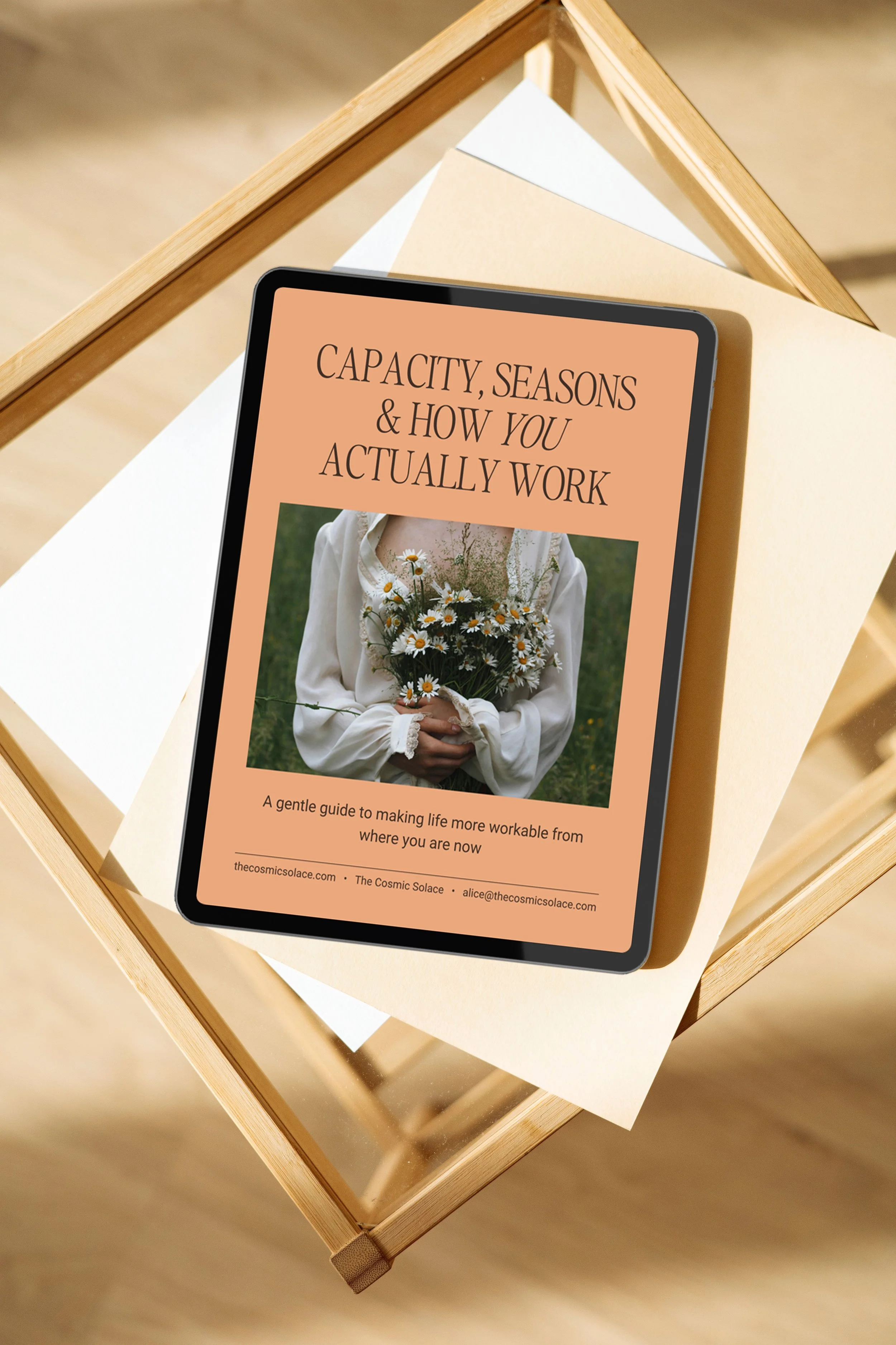 A tablet computer on a glass top table displaying a book or guide titled "Capacity, Seasons & How You Actually Work" with a woman holding a bouquet of daisies, dressed in white, on the cover.