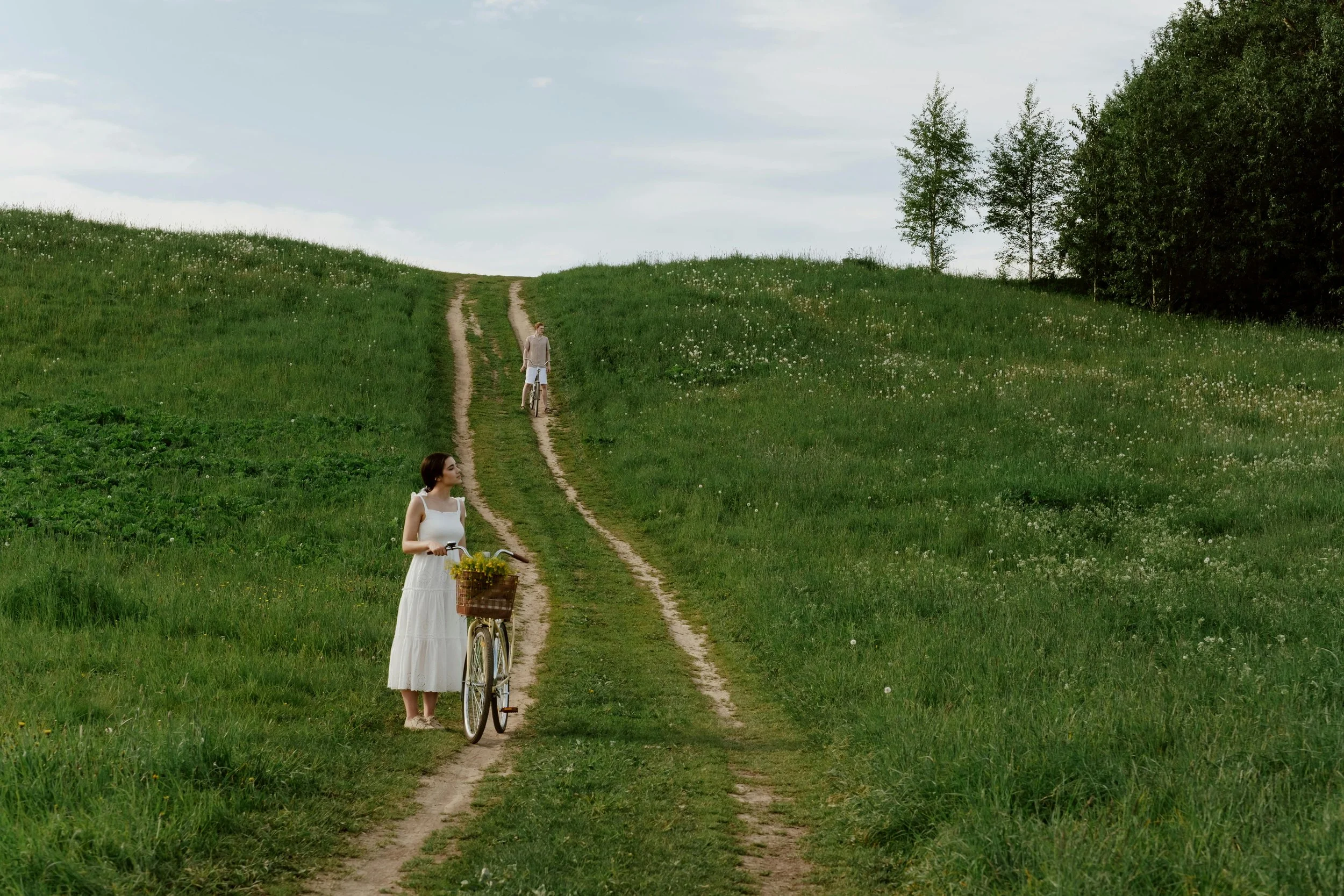 A woman in a white dress holding a bicycle with a basket of yellow flowers stands on a grassy dirt path in a green field with rolling hills. A man with a bicycle further up the path is visible, with trees in the background under a partly cloudy sky.