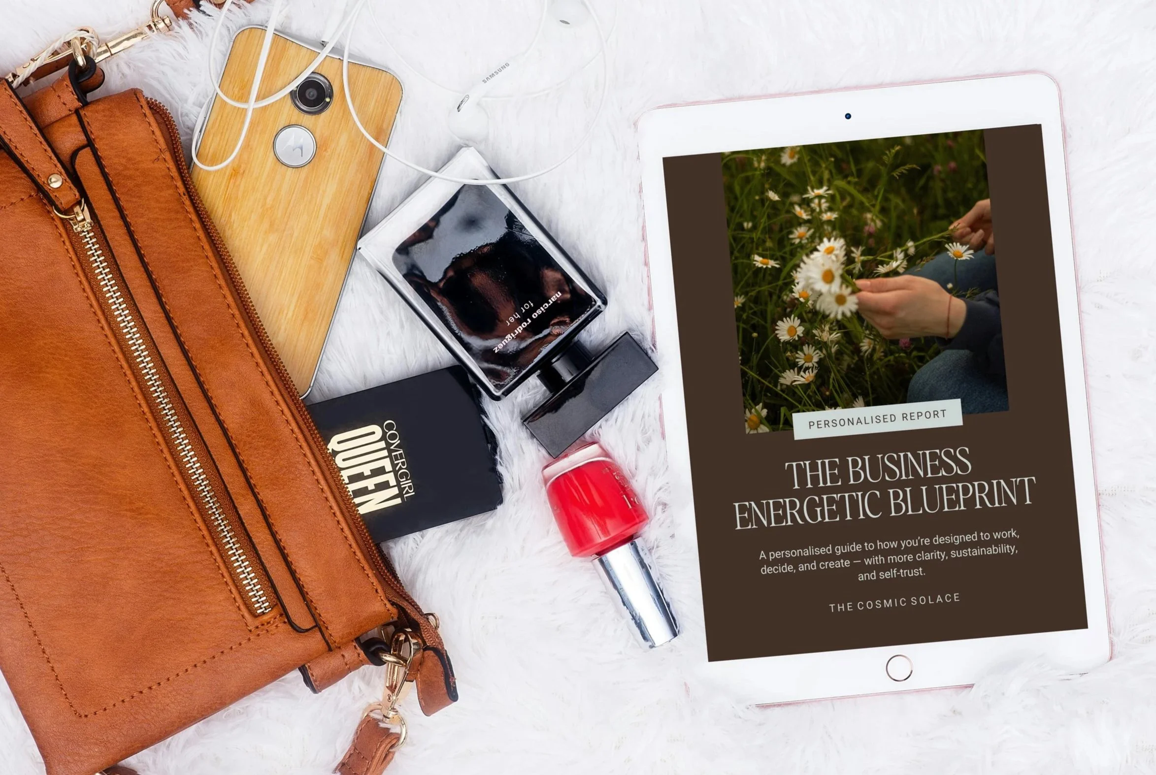 A flat lay of various items on a white furry surface, including a brown leather bag, a smartphone with white earbuds, a perfume bottle, a red lipstick, a black package labeled "CoverGirl Queen," and an iPad displaying a report titled "The Business Energetic Blueprint" with a photo of daisies.