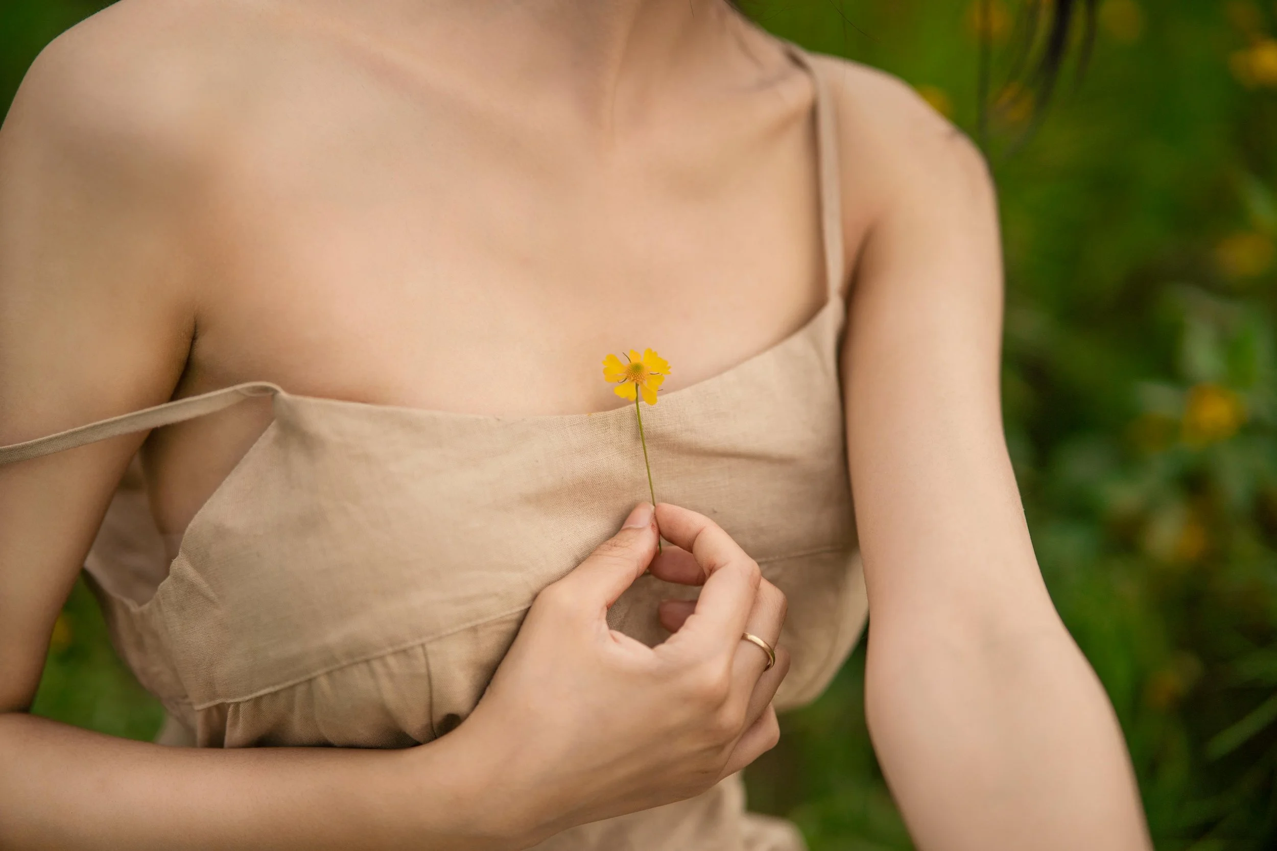 Person in a neutral-toned dress holding a small yellow flower against their chest in a green outdoor setting.