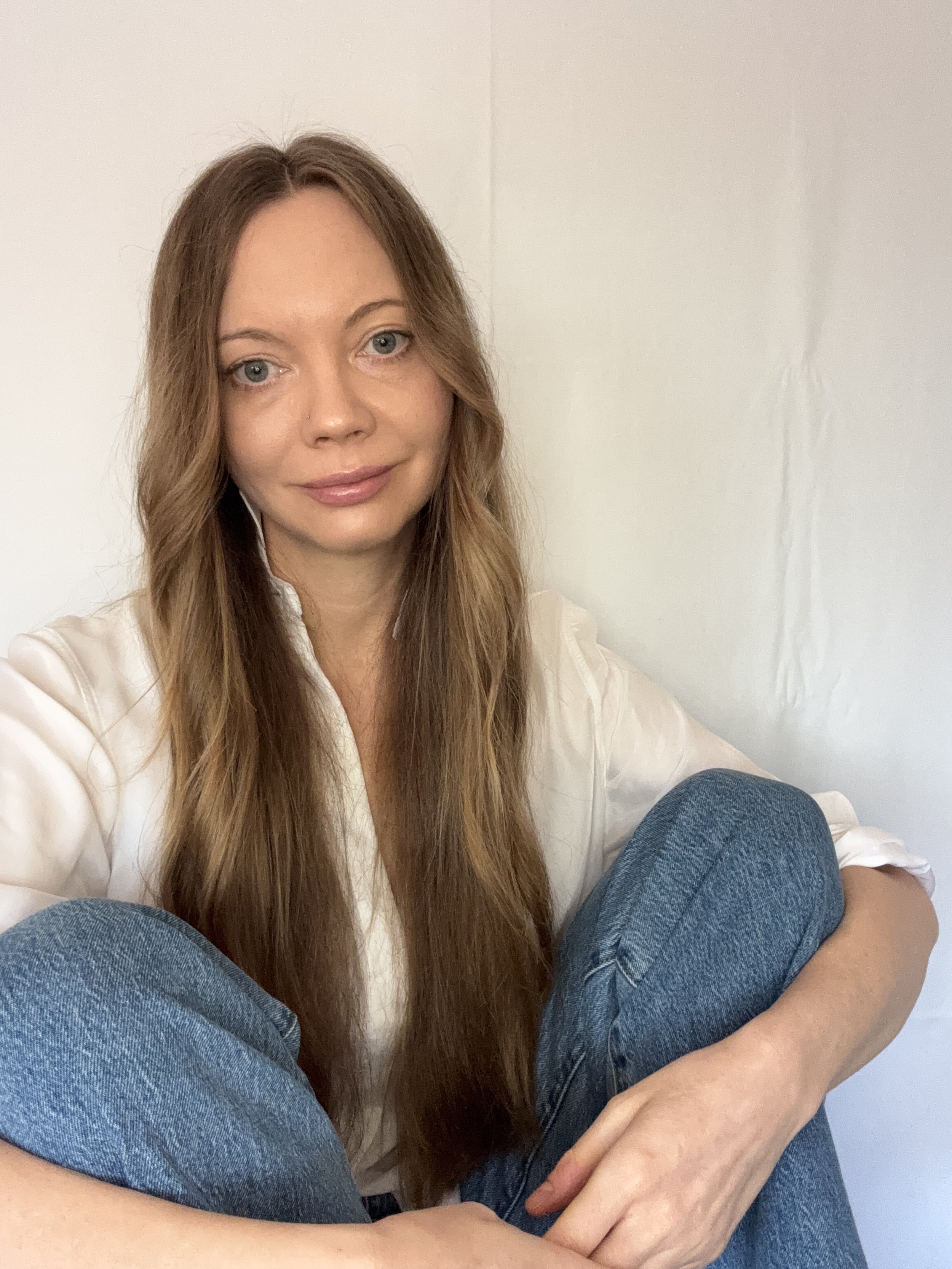 A woman with long, wavy brown hair sitting on the floor with her knees up, wearing a white shirt and blue jeans, against a plain white wall.