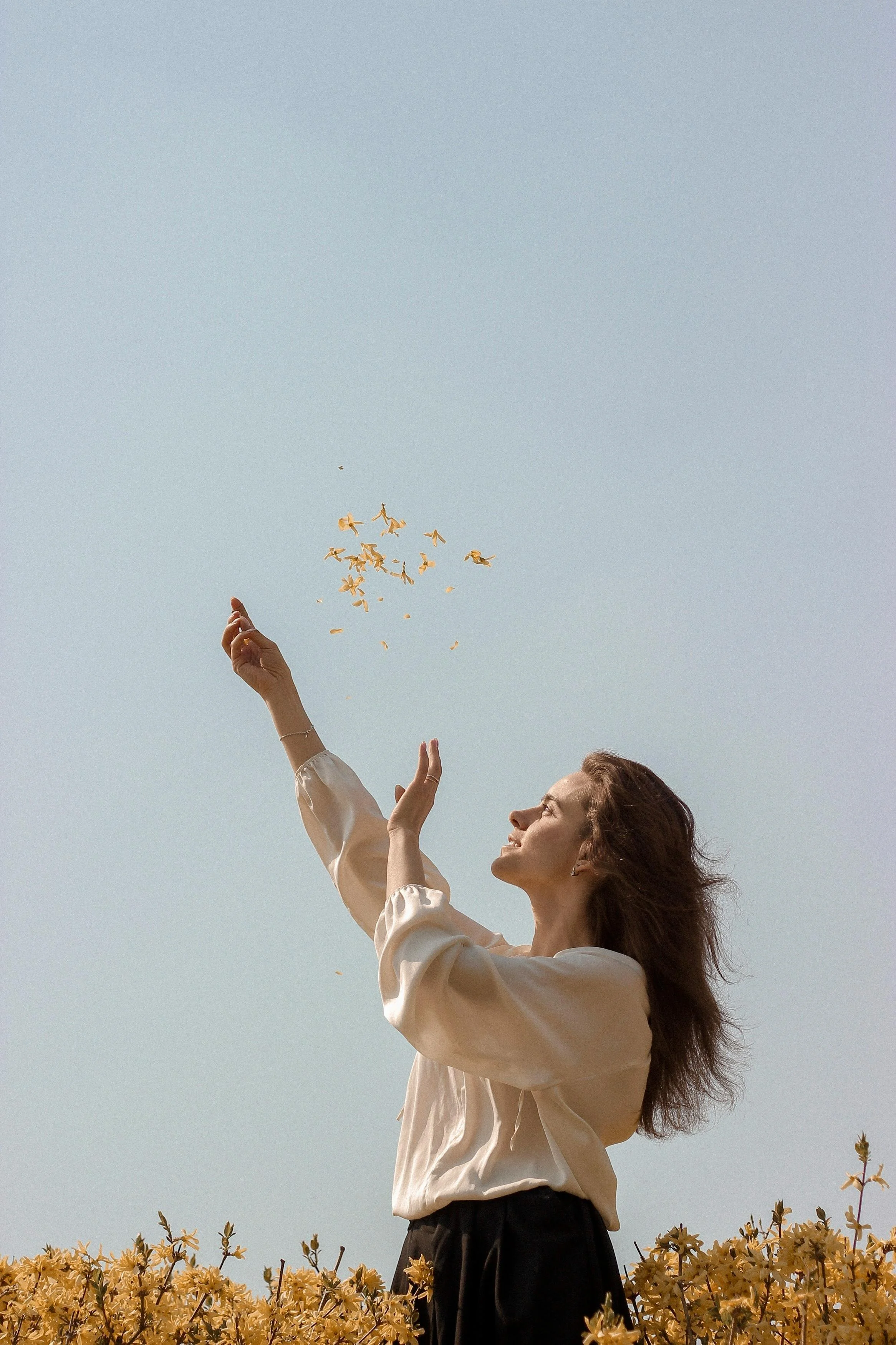 A woman standing in a field of yellow flowers, throwing flower petals into the air, smiling and facing the sun on a clear day.