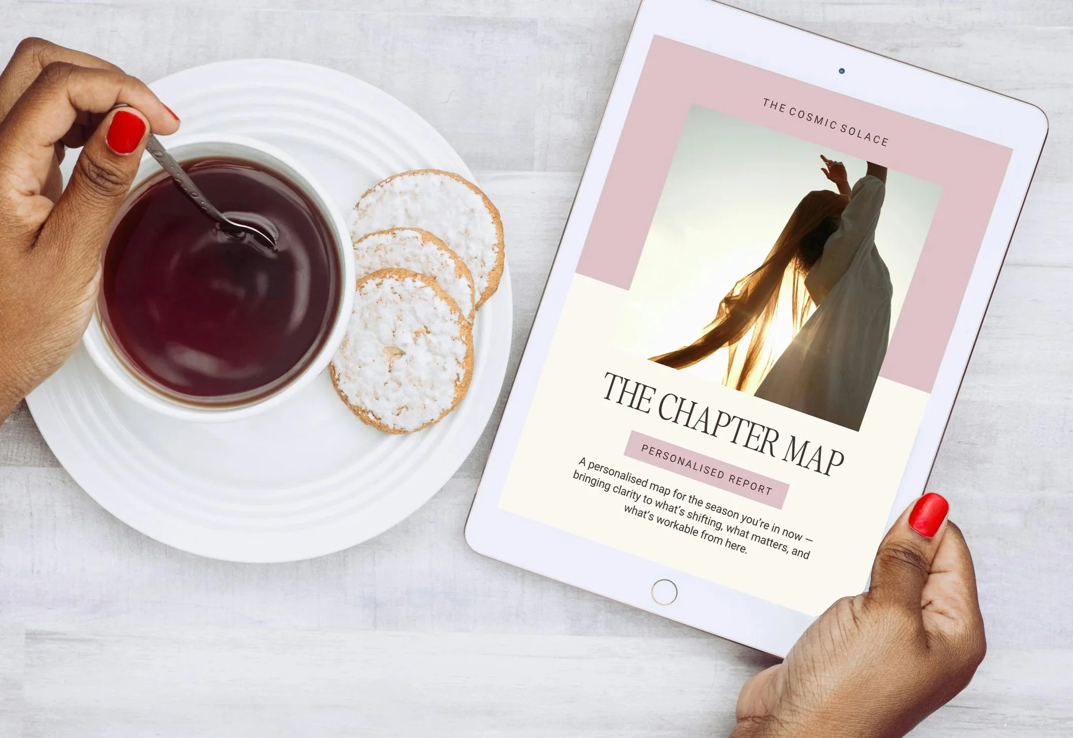 Hand with red nail polish holding a tablet displaying a book cover titled 'The Chapter Map' with an image of a woman in sunlight. Beside the hand, there is a white plate with three coconut cookies and a cup of dark tea on a light-colored wooden surface.