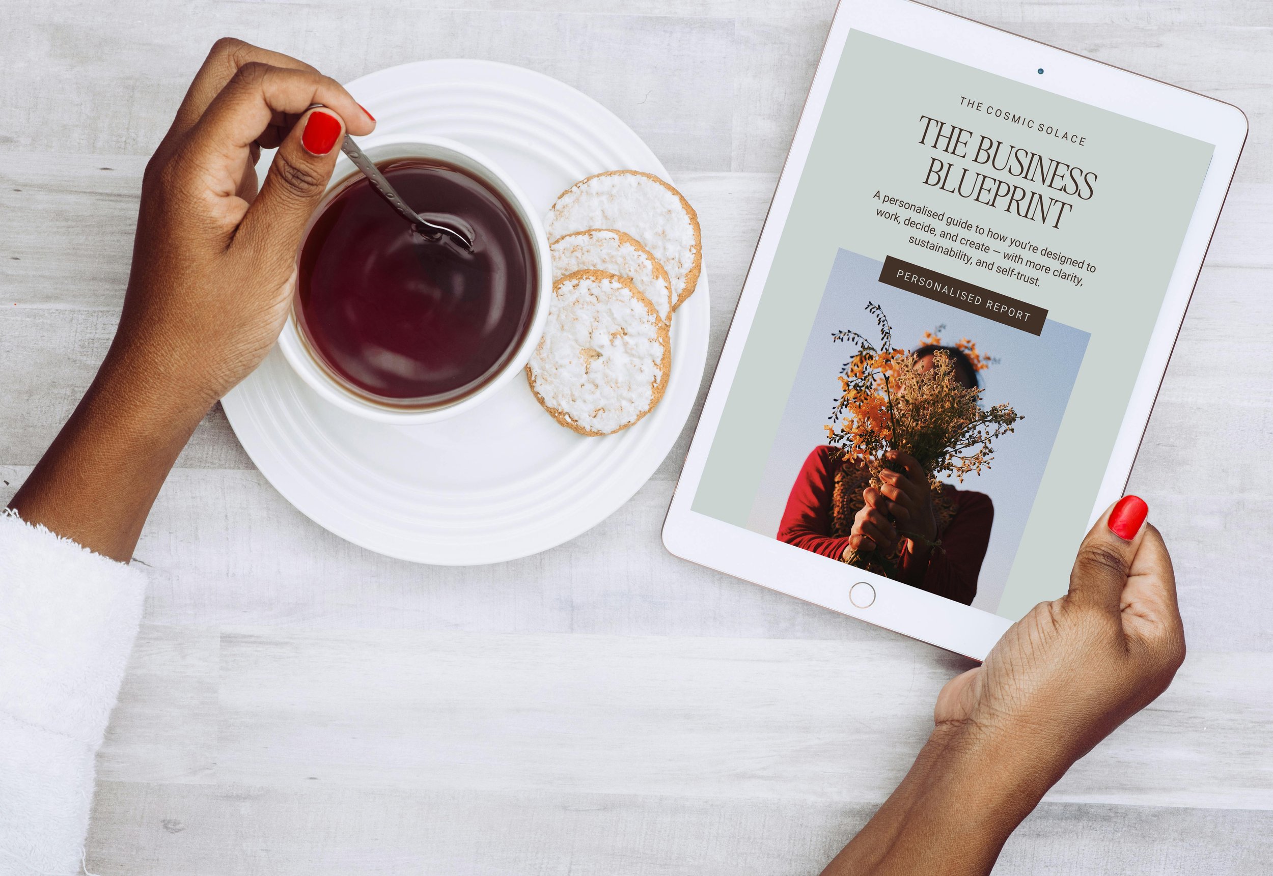 A person's hand with red painted nails is holding a tablet displaying a book titled 'The Business Blueprint' alongside a cup of tea and some cookies on a white plate, on a light-colored wooden surface.