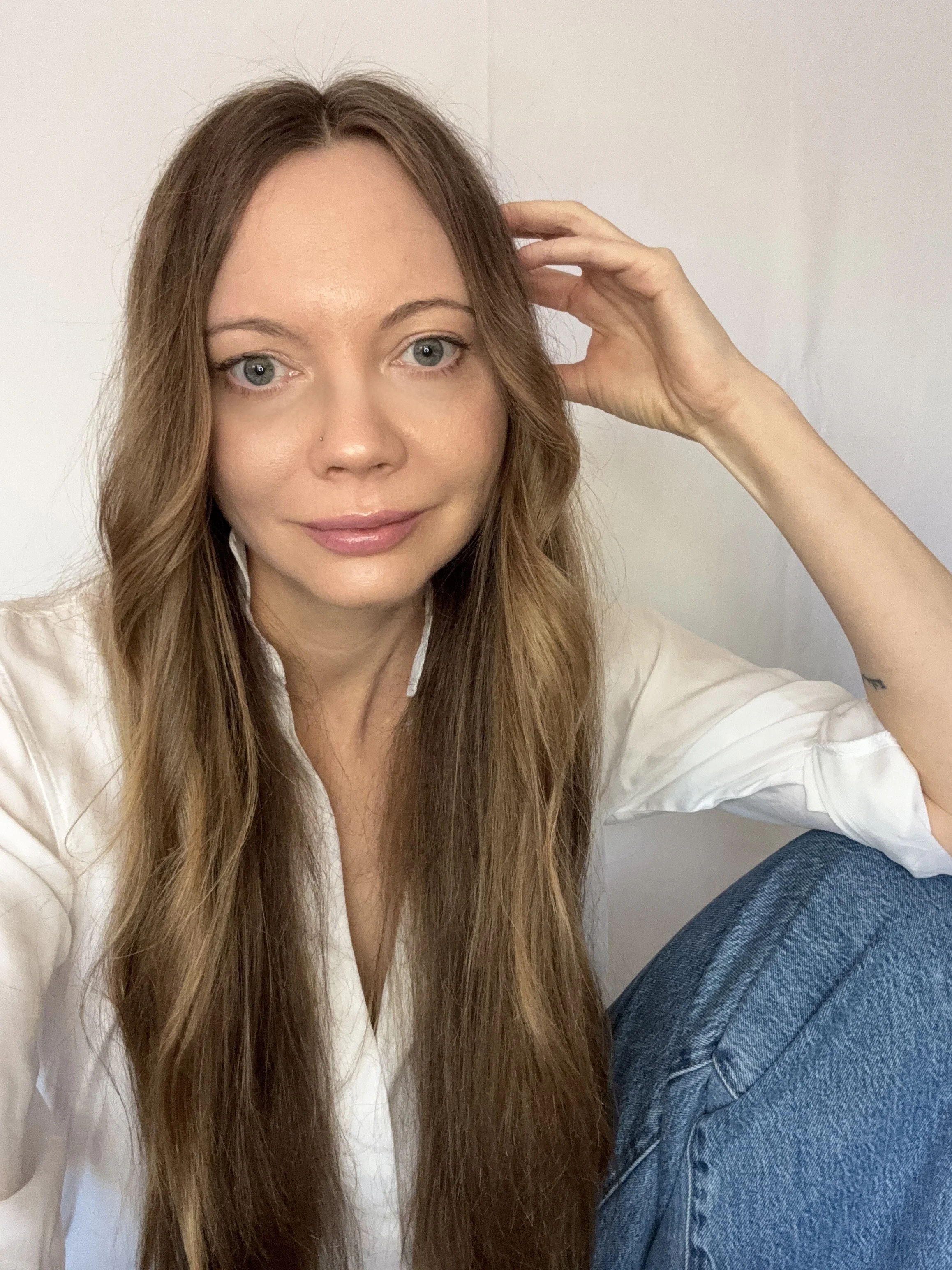 A woman with long, wavy brown hair wearing a white shirt, sitting against a plain white background, looking at the camera with a slight smile.