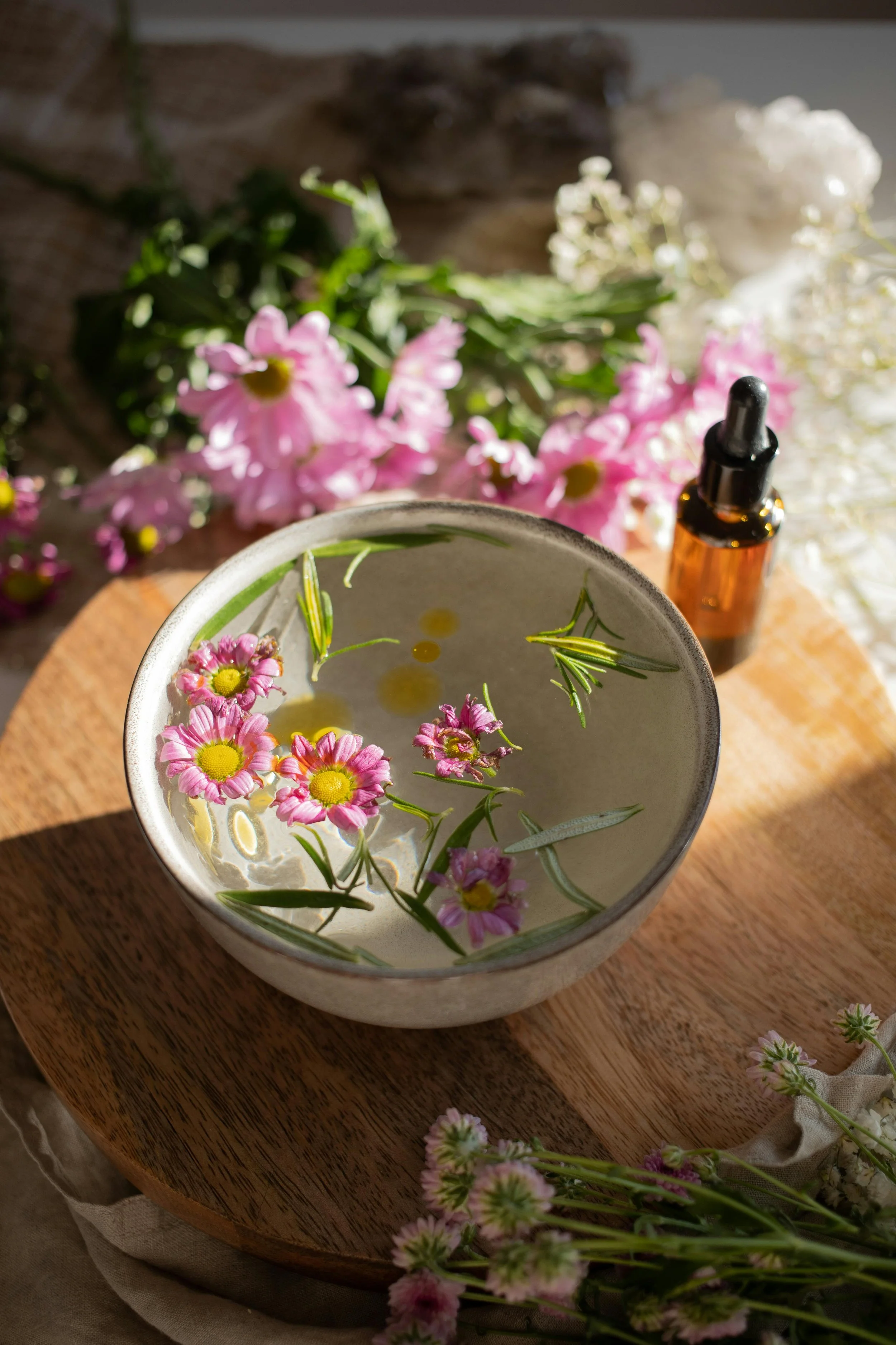 A bowl filled with water and floating pink flowers and green leaves, placed on a wooden surface surrounded by additional flowers and a small brown bottle with a dropper.