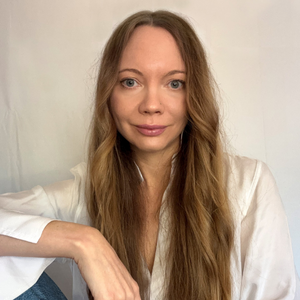 A woman with long, wavy blonde hair wearing a white shirt, sitting indoors against a plain white wall.