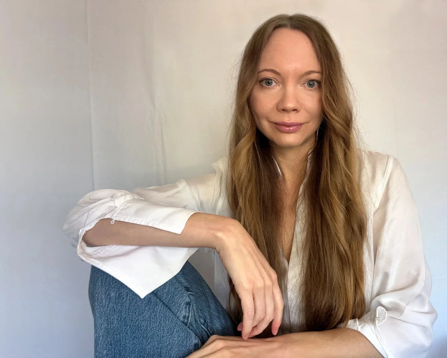 A young woman with long, wavy, reddish-brown hair sitting on the floor, wearing a white satin blouse and blue jeans, against a plain white wall.