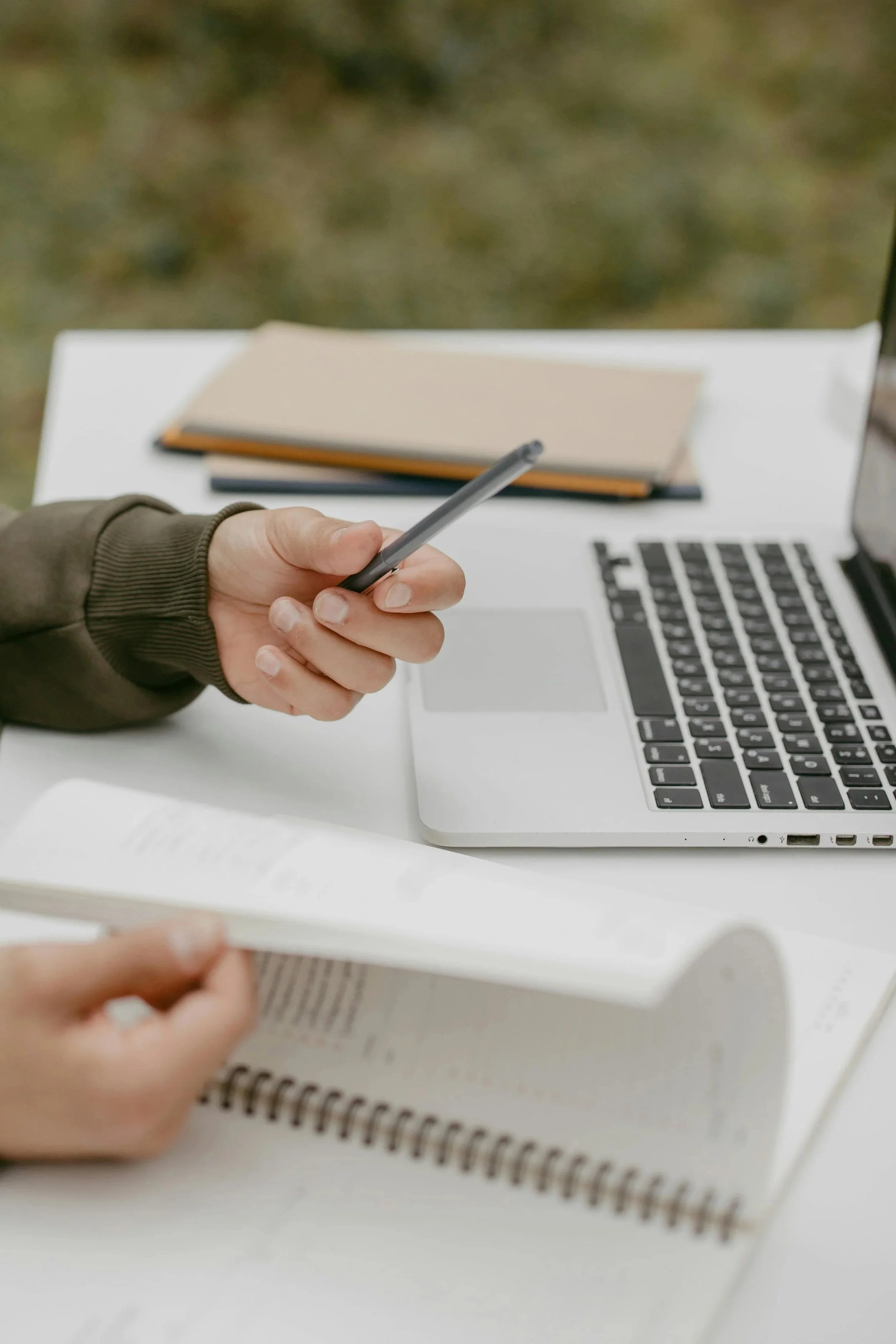 Person holding a smartphone over a white desk with a laptop, notebooks, and a book.