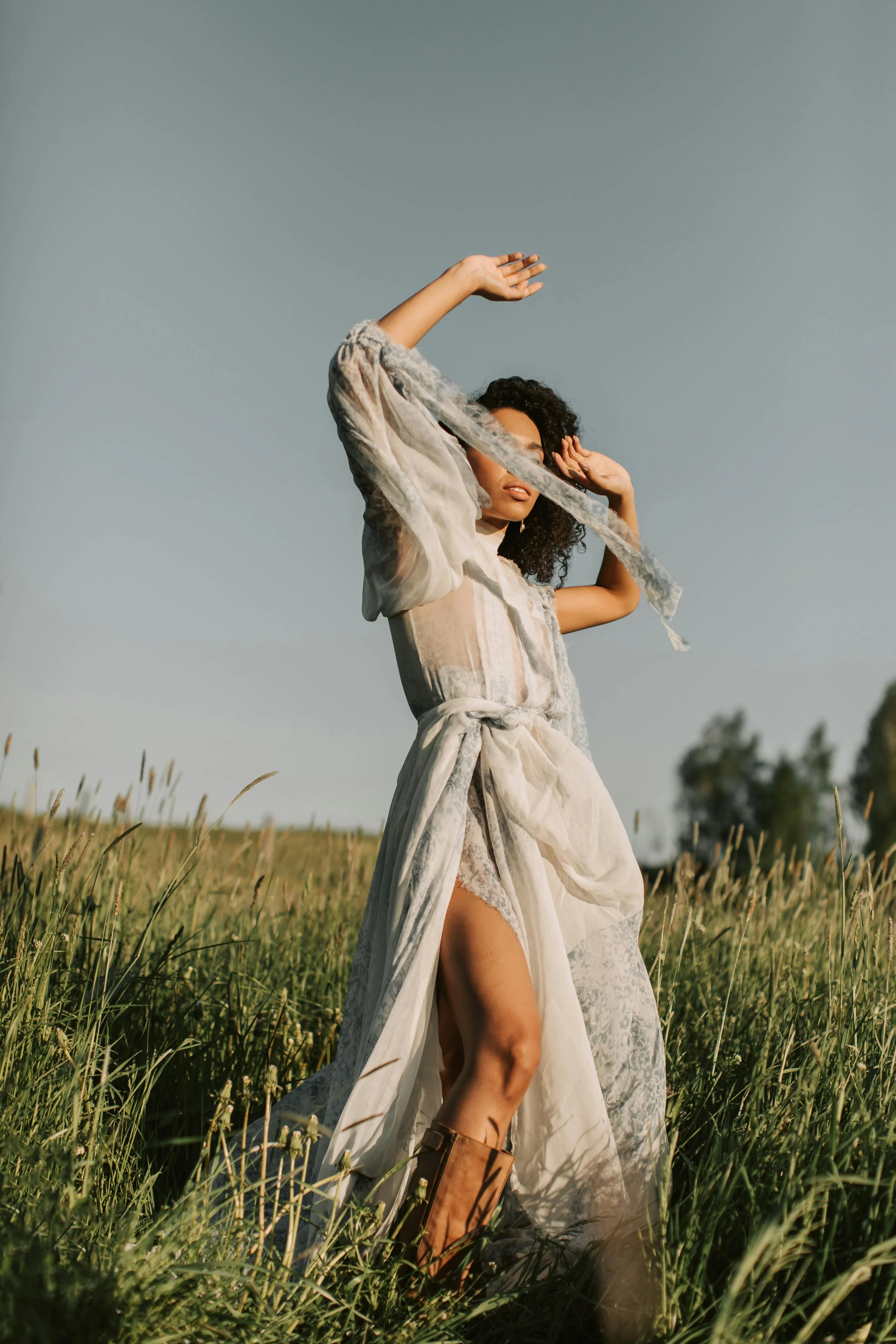 Woman in a flowing dress and boots standing in a grassy field under a clear sky, with her hands raised near her head.