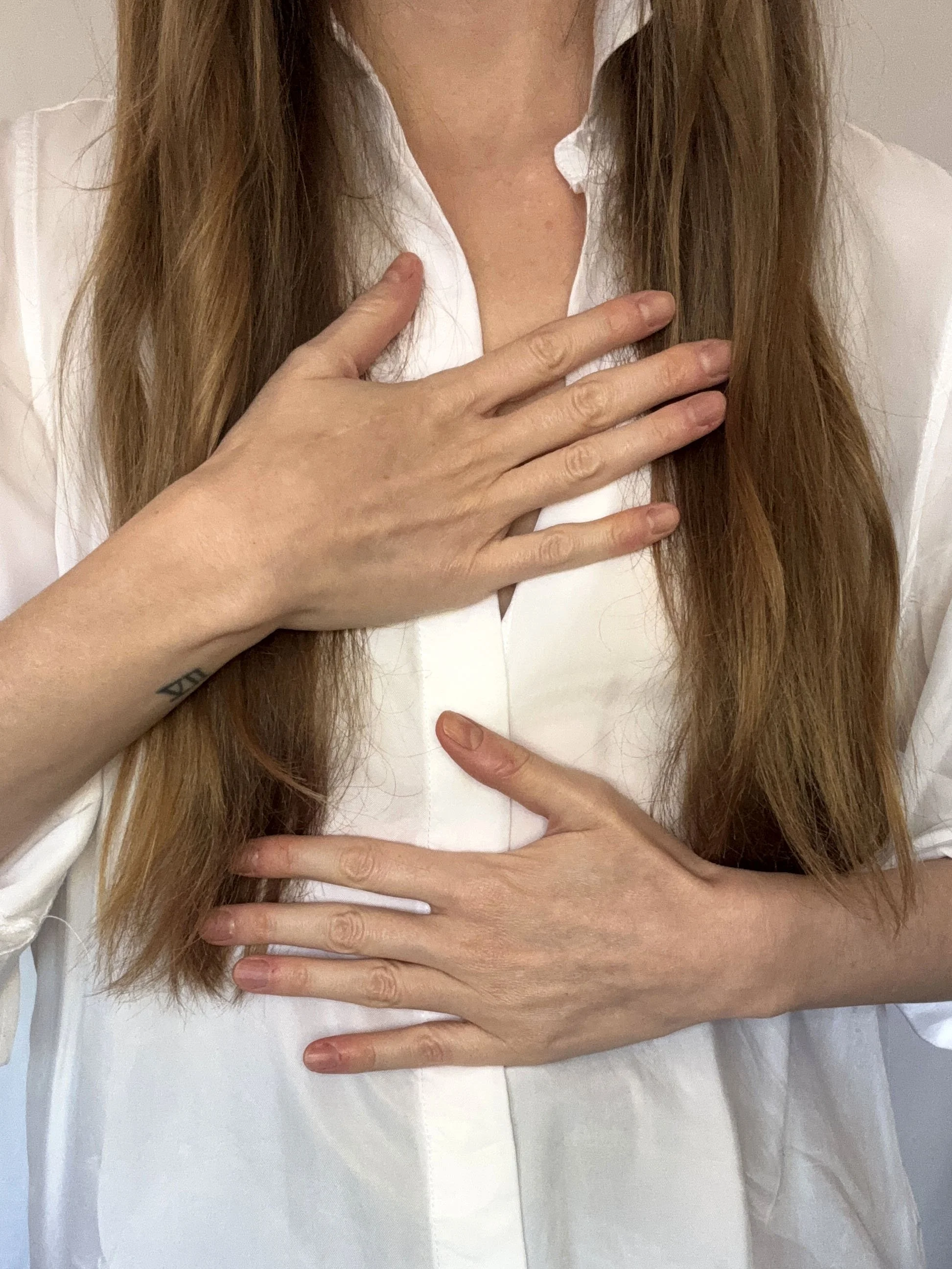 A woman with long auburn hair wearing a partially open white blouse, holding one hand over her heart center with fingers spread apart, and another hand at the solar plexus - stomach area, fingers spread apart.