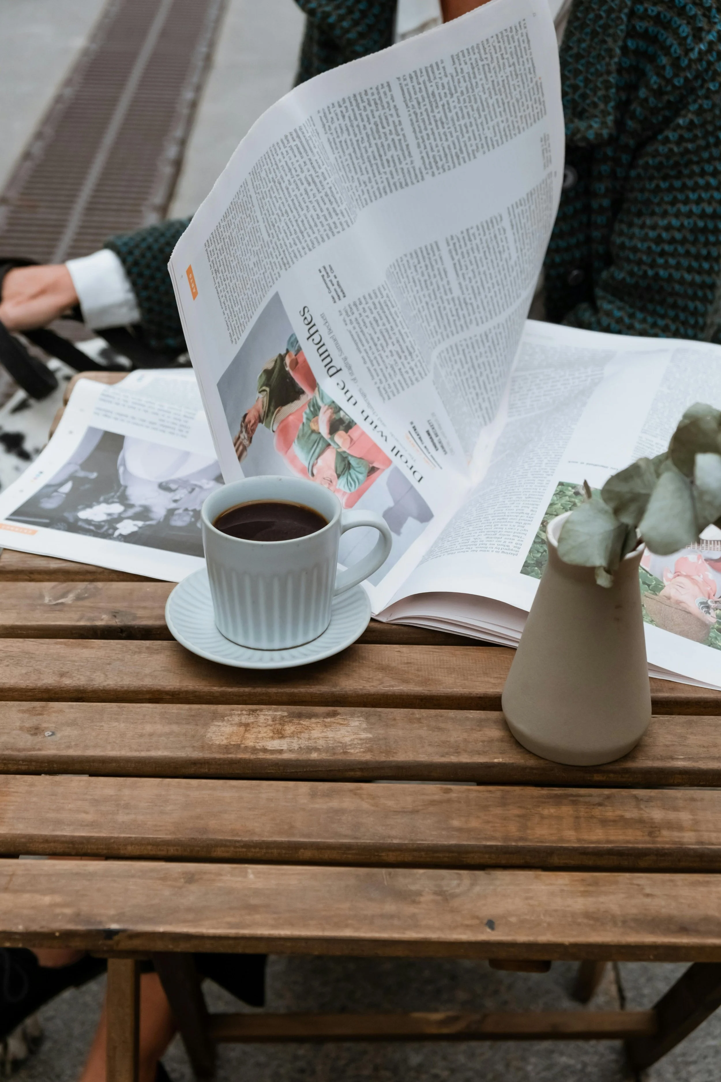 A wooden table with a white cup of coffee, an open newspaper, and a beige vase with dried flowers, outdoors with a person sitting in the background.