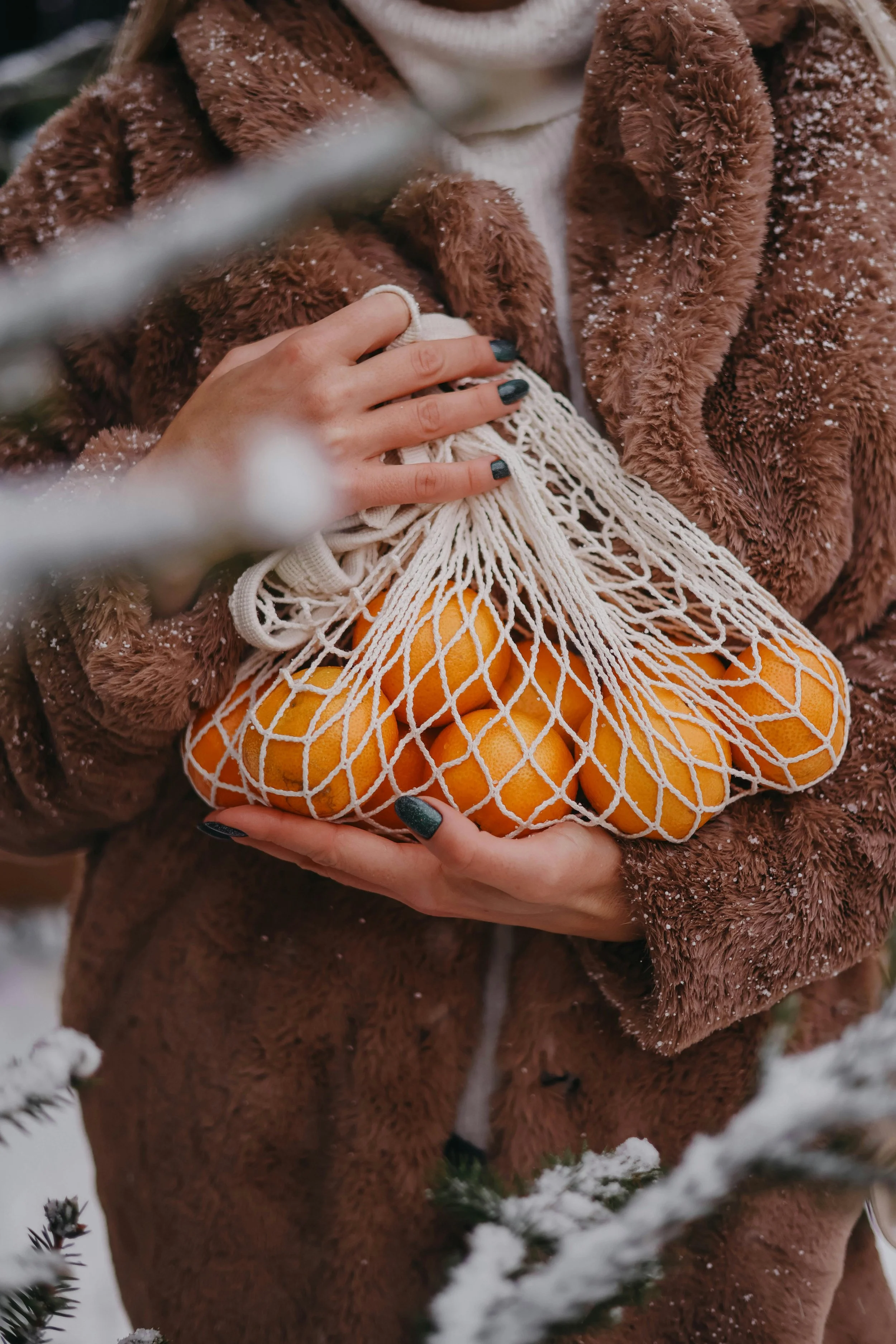 A woman in a warm furry brown jacket, holding a mesh bag of oranges outdoors in winter, with snow-covered branches in the foreground.