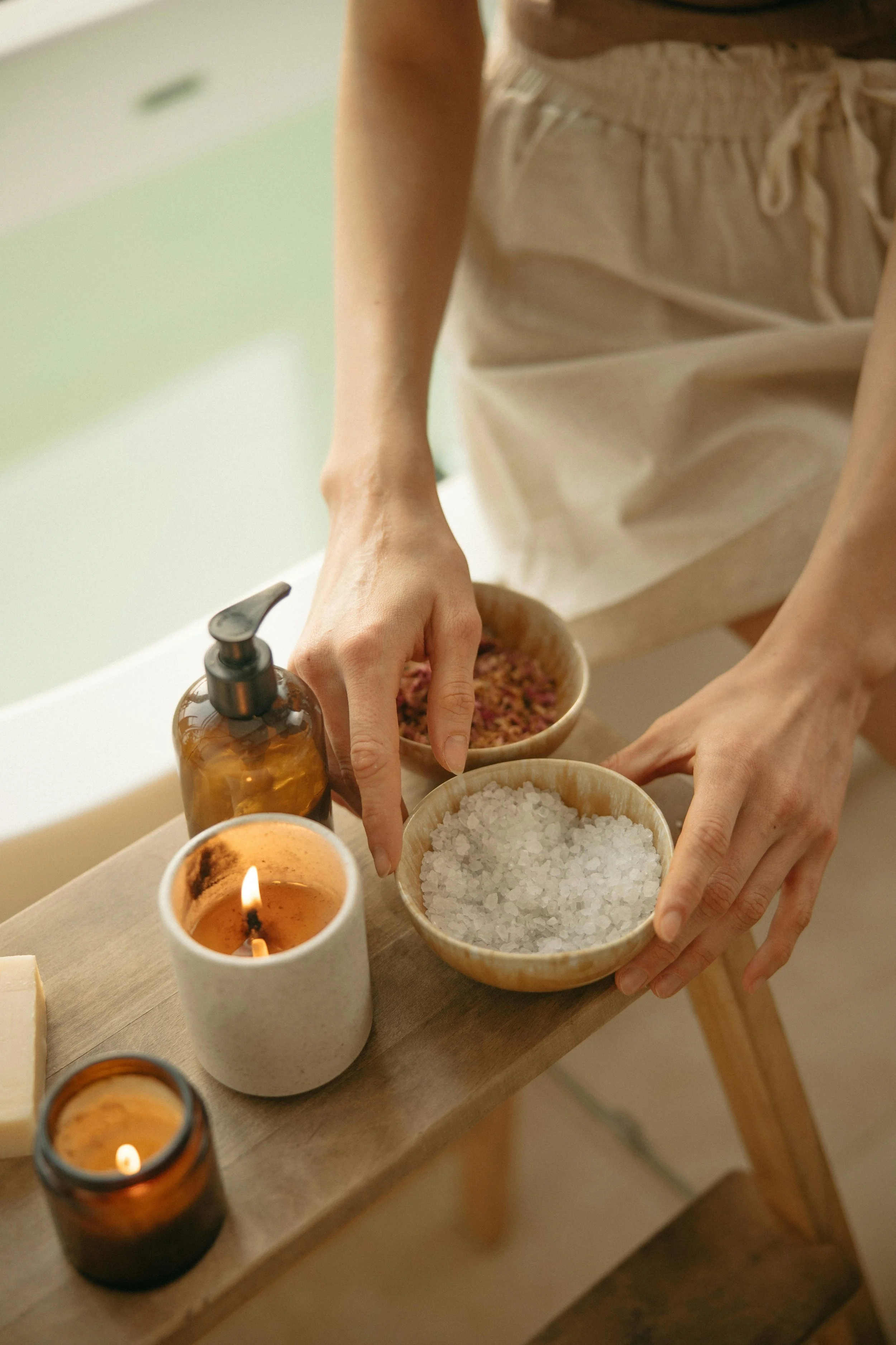 A person in a light-colored apron arranging bowls of bath salts and dried herbs on a wooden table, at the edge of a bathtub, with lit candles nearby.