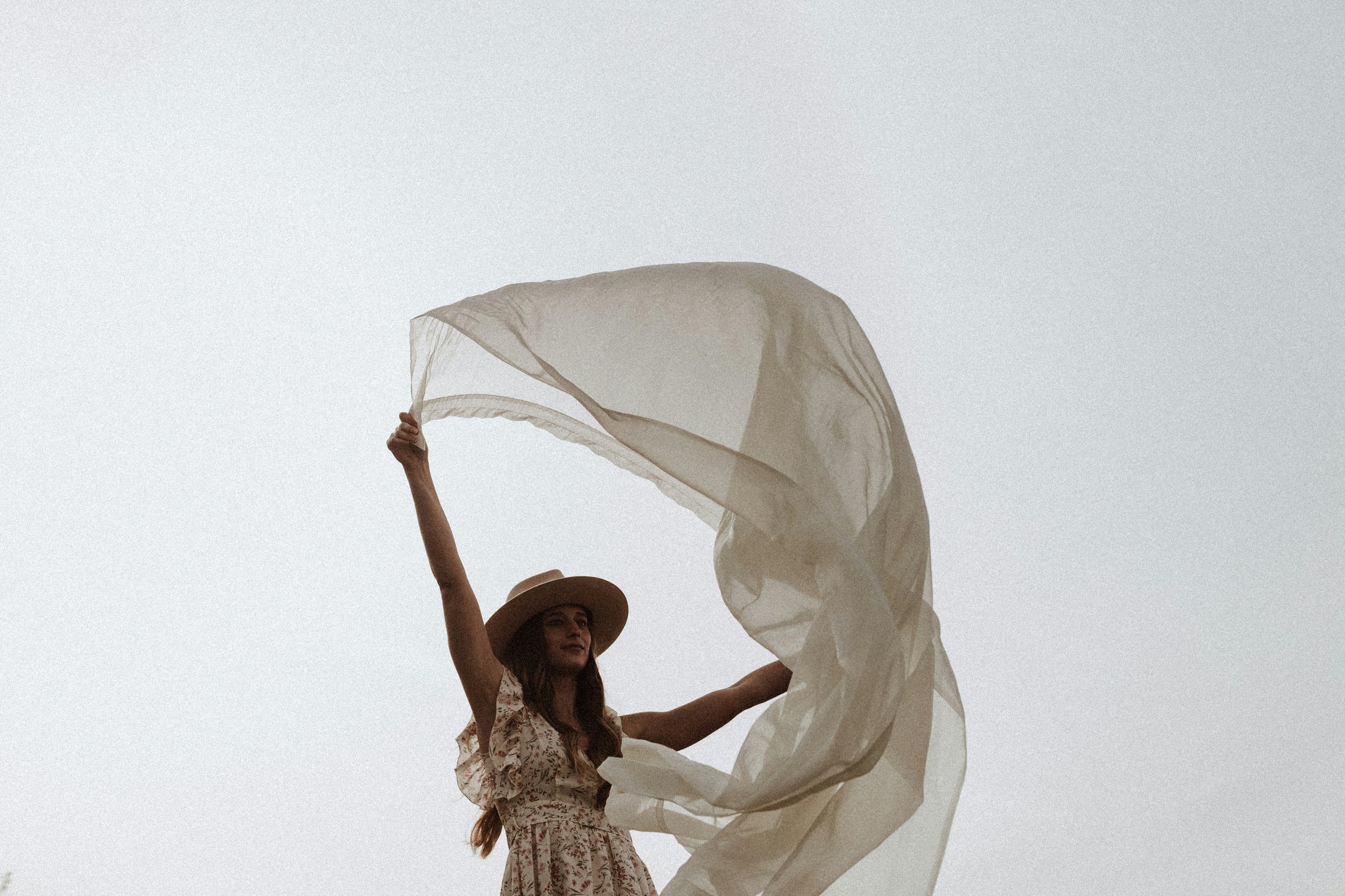 Woman wearing a hat holding a large, flowing white fabric against a light sky.