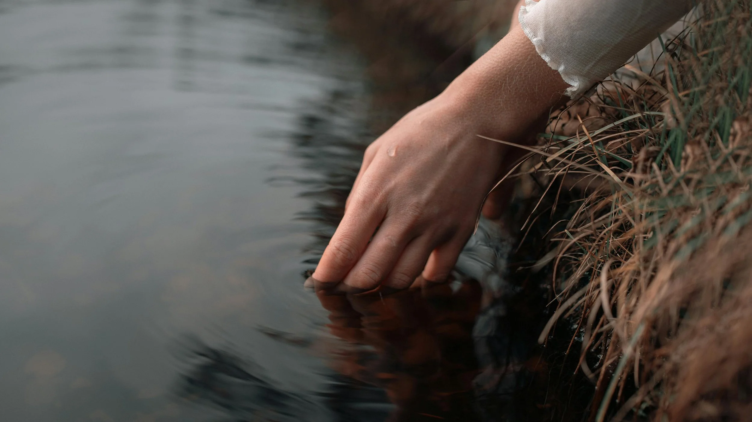 Close-up of a hand reaching down to touch the surface of still water at the water's edge