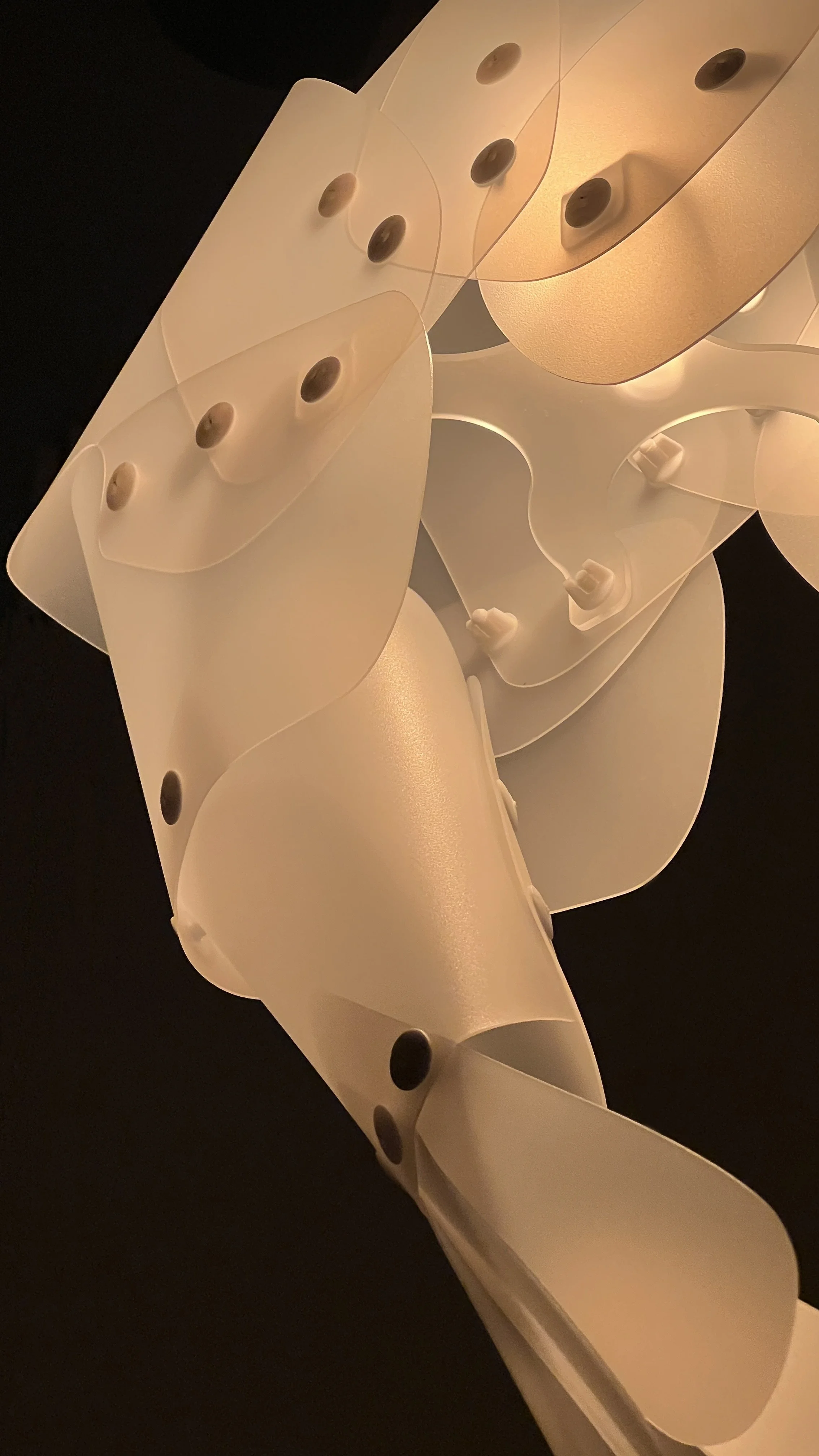Close-up of a modern ceiling light fixture with white curved panels and small dark circular accents, illuminated against a dark background.