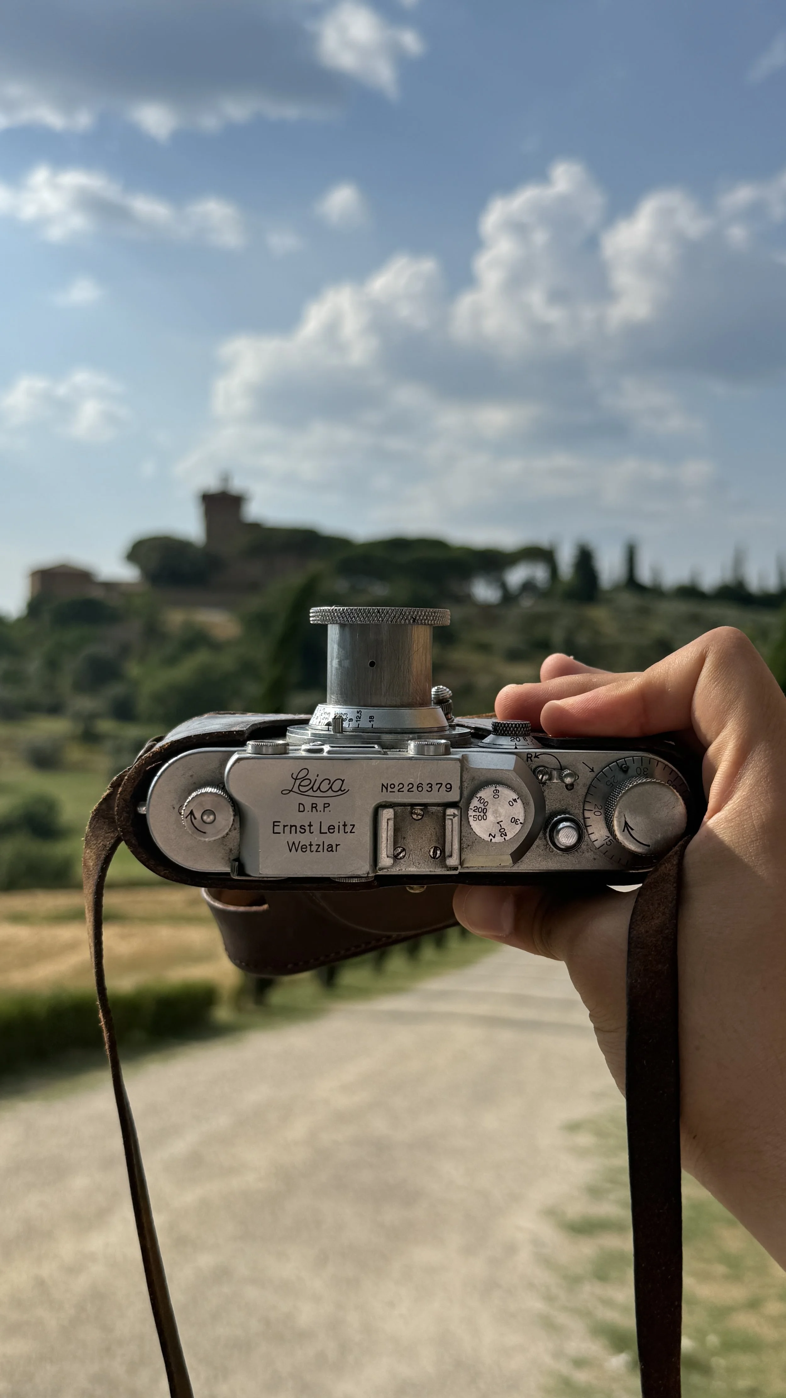 A vintage Leica camera held up by a hand in a scenic outdoor setting, with a historical building and cloudy sky in the background.