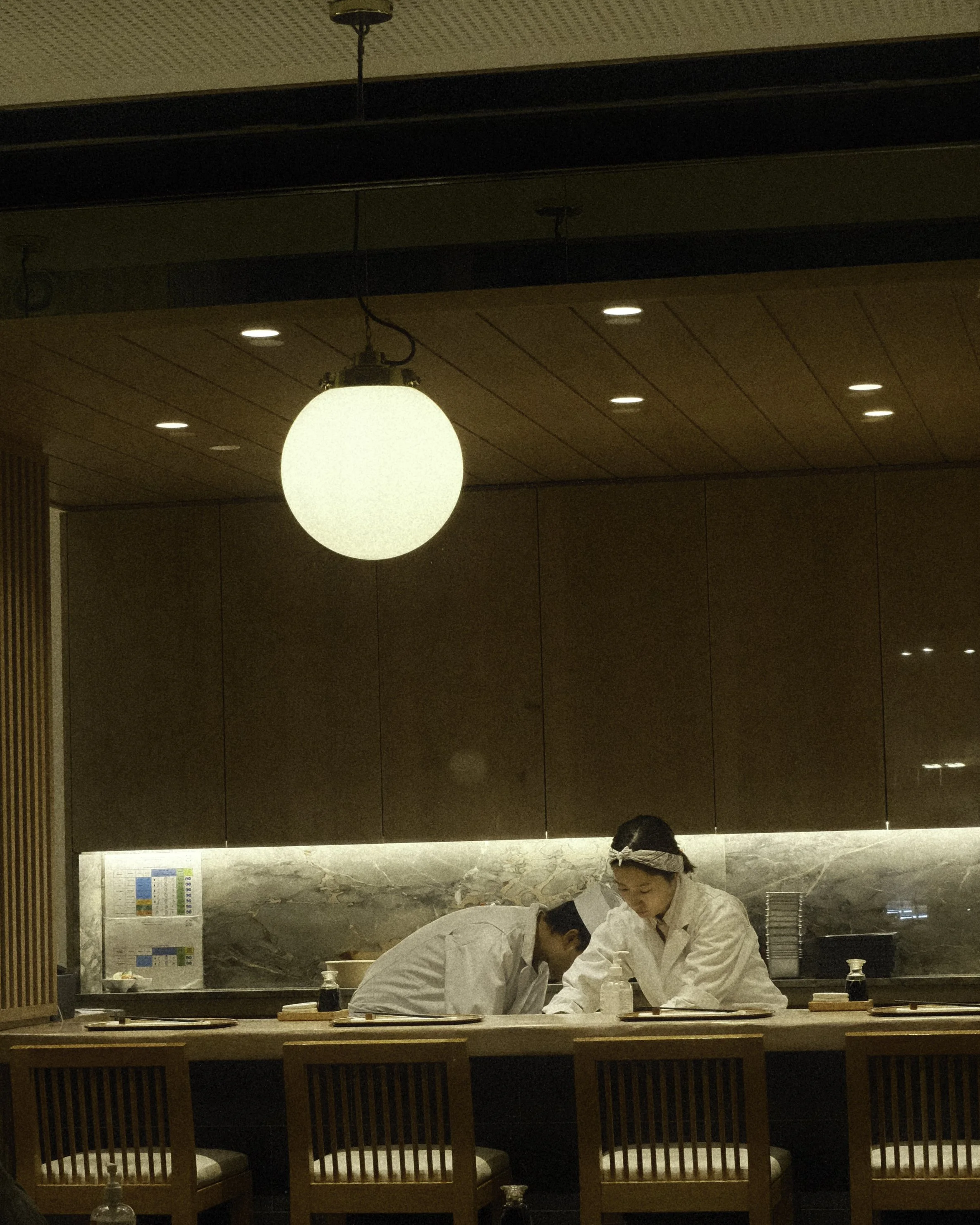 Two chefs in white uniforms and chef hats working behind a kitchen counter in a restaurant, with warm lighting and wooden decor.