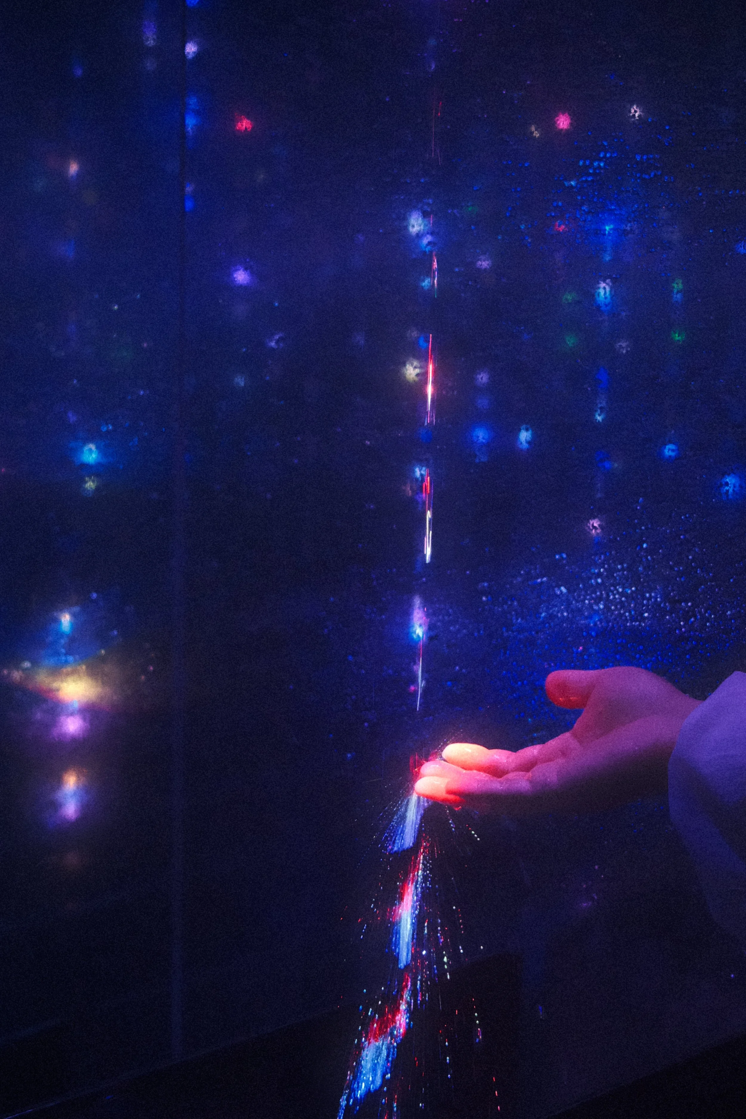 A person's hand extends toward a display of colorful, sparkling light projections resembling a starry night or galaxy.