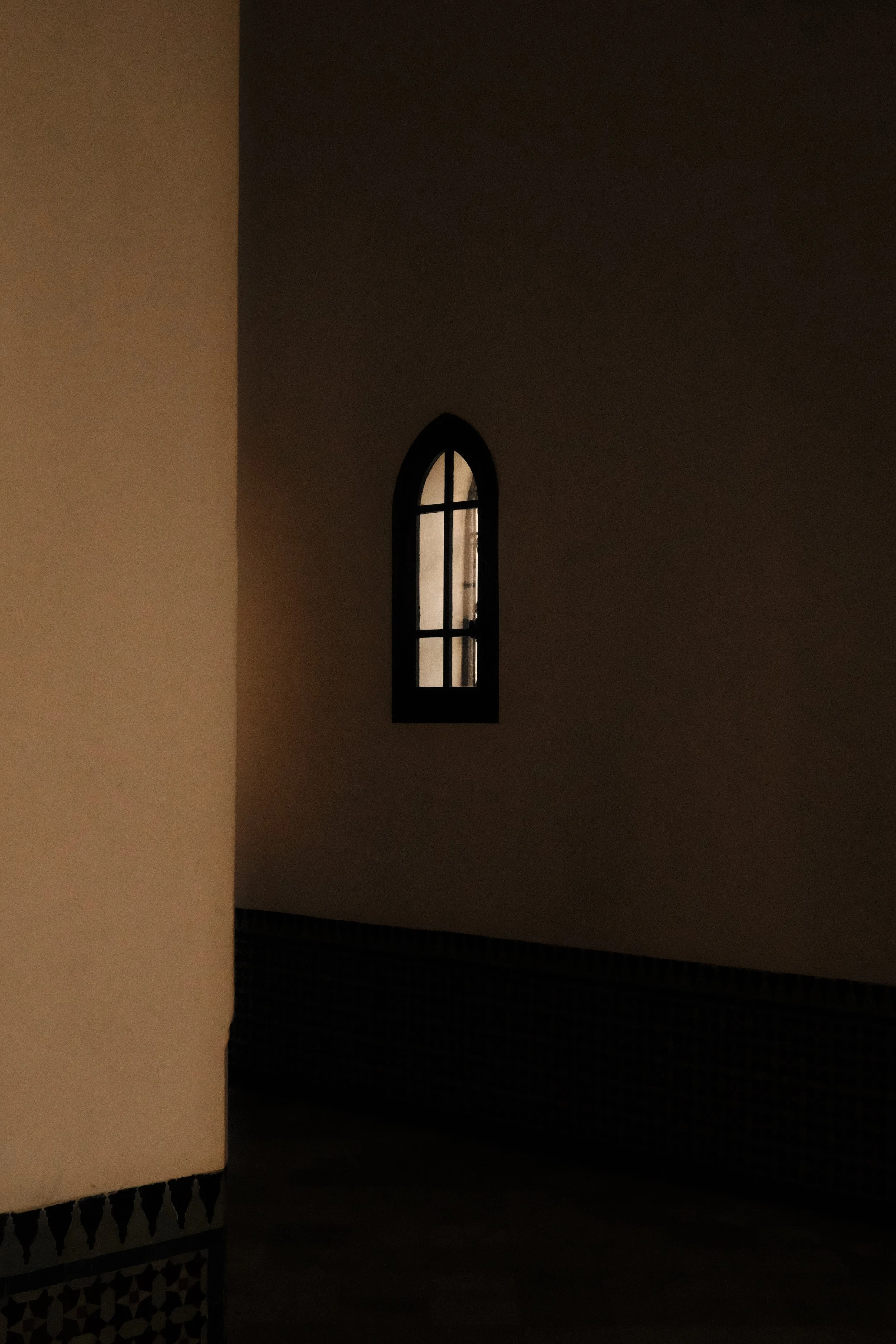 A dark interior with a tall, narrow, arched window with black framing, showing light coming in from outside. The walls are beige, and there's a patterned border near the floor.