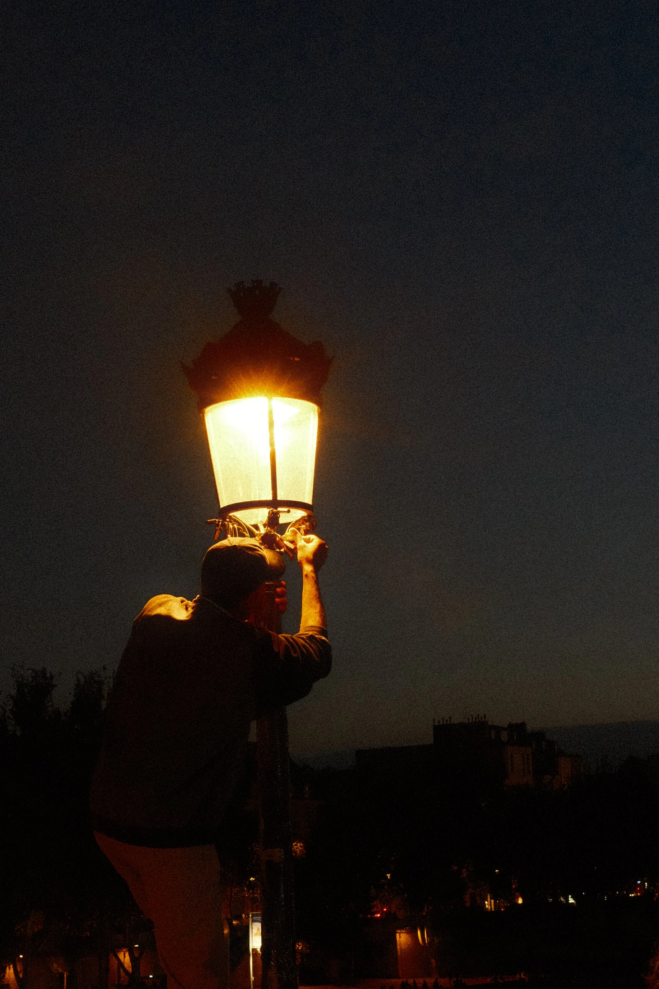 Person inspecting or repairing a street lamp at night under a dark sky with buildings in the background.