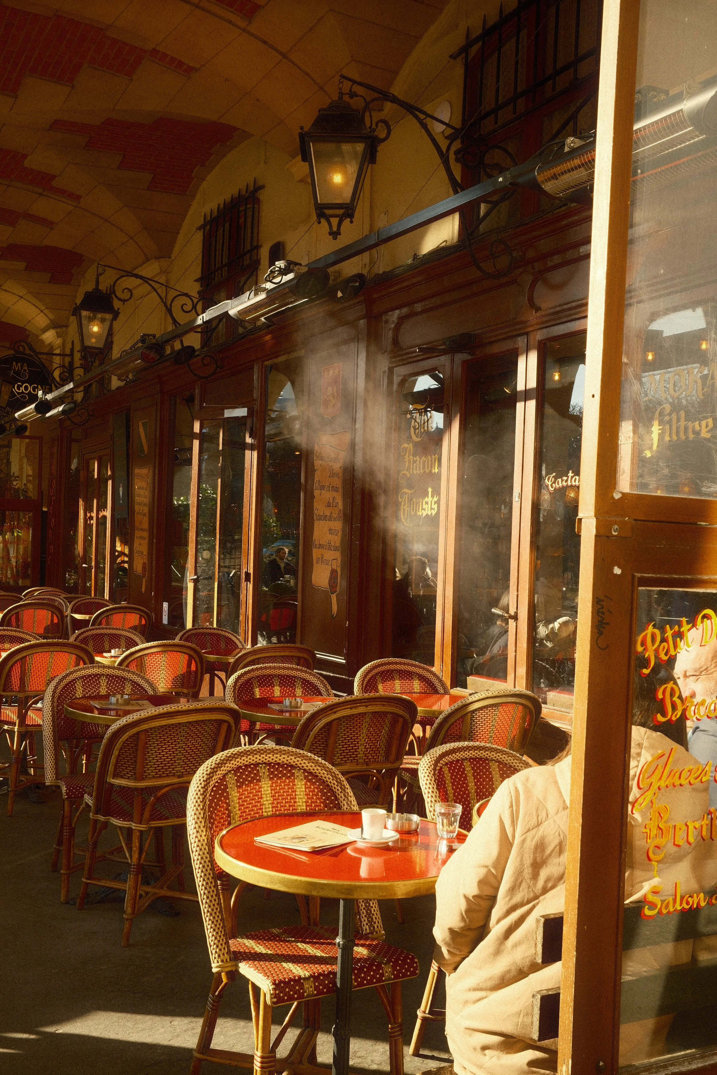 Outdoor café with red tables and chairs, some with cups and glasses, under an arched ceiling, with customers sitting outside.