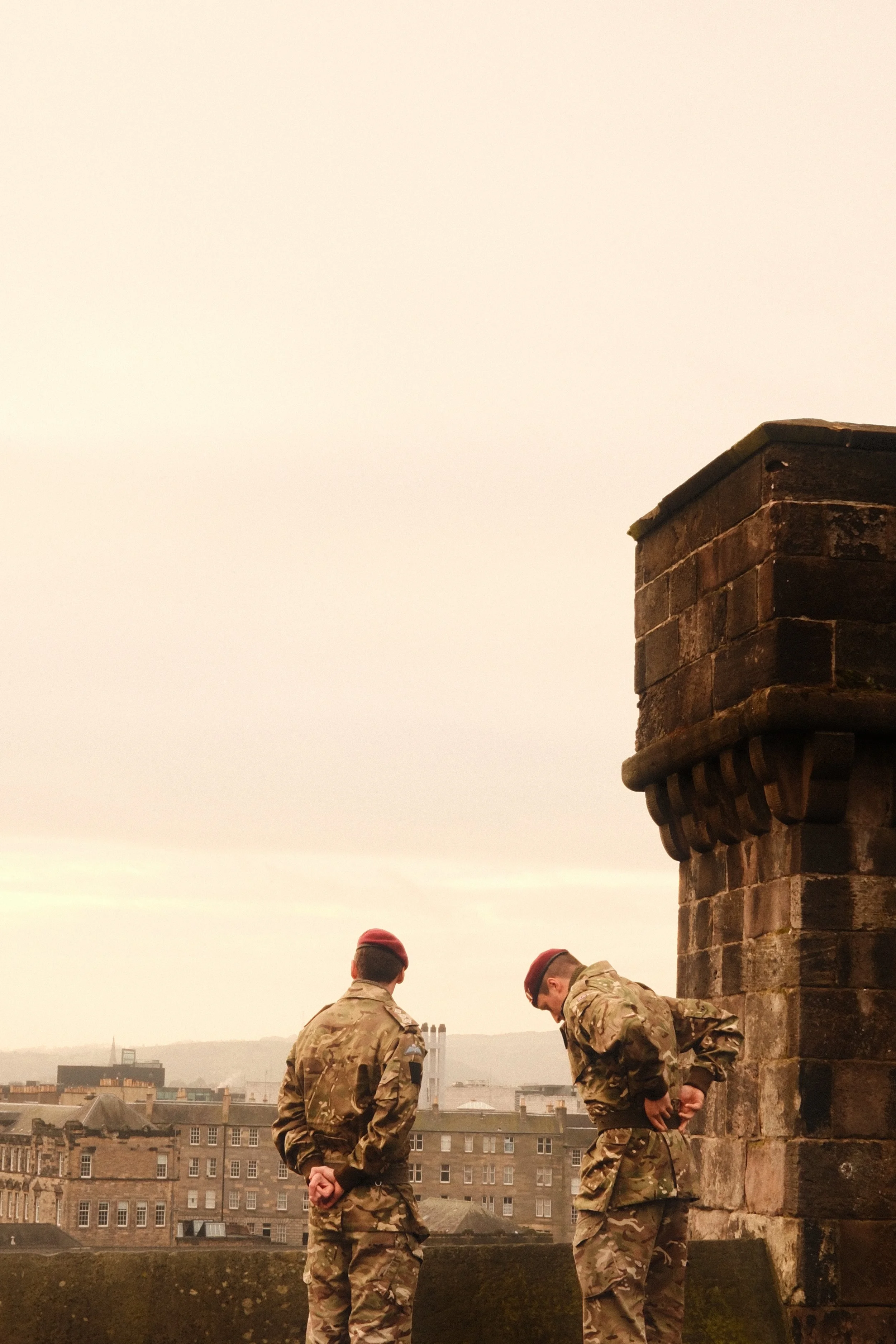 Two soldiers in camouflage uniforms and red berets stand outdoors on a cloudy day near a stone structure, with city buildings in the background.