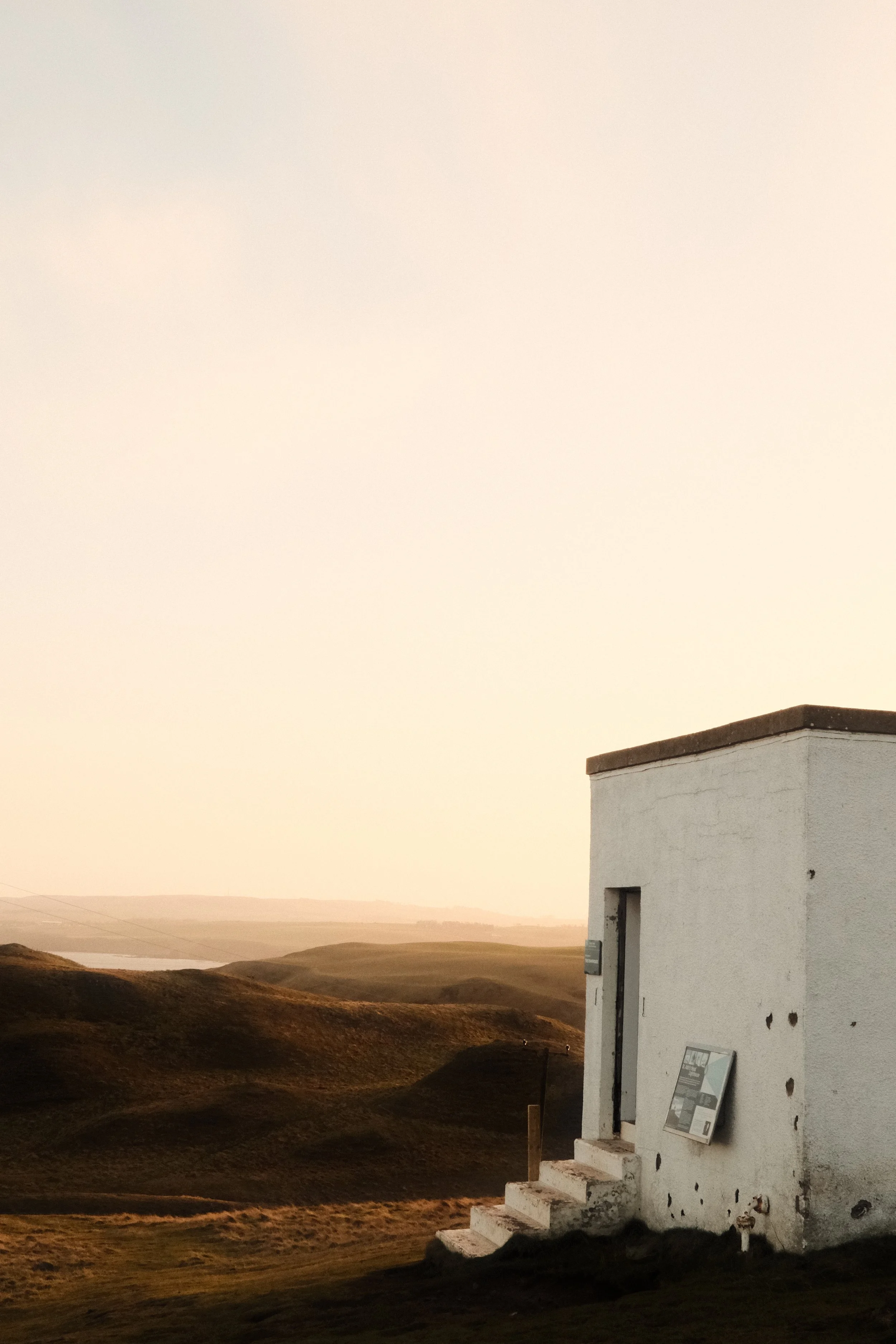 A small white building with steps leading up to the door, set against rolling hills at sunset.
