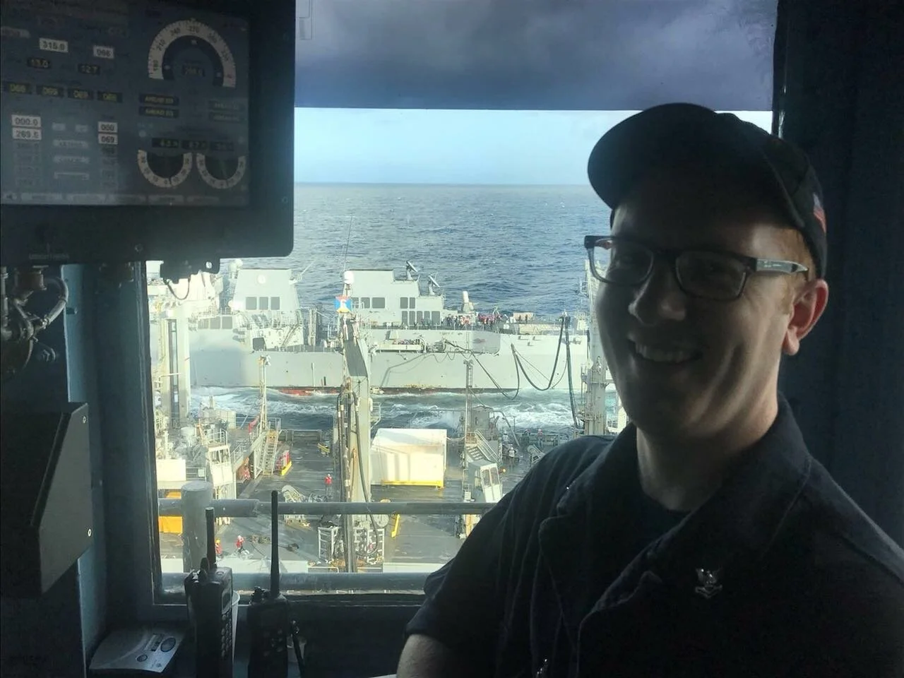 Person smiling, wearing a cap and glasses, inside a ship's control room with a view of a naval vessel on the ocean through the window.
