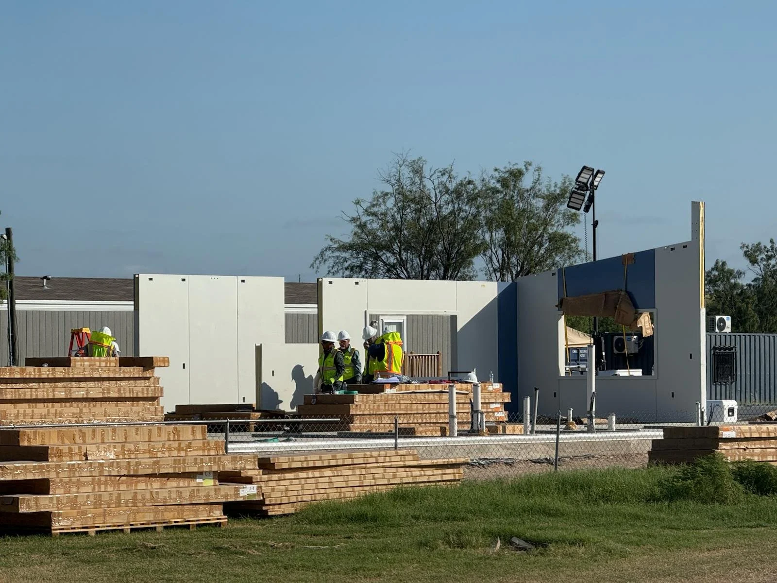 Construction site with workers wearing safety gear, surrounded by stacks of wood and a partially built wall. Bright daylight, clear sky, and some trees in the background.