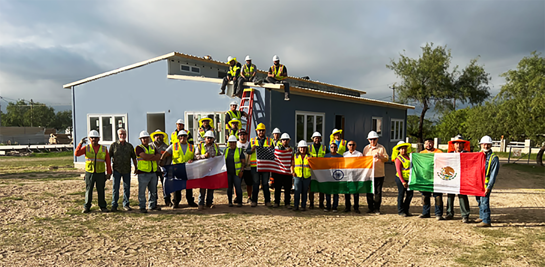 Group of construction workers and officials standing in front of a house under construction, holding flags of Texas, India, and Mexico, with some workers sitting on the roof and others standing on the ground, wearing safety helmets and vests.