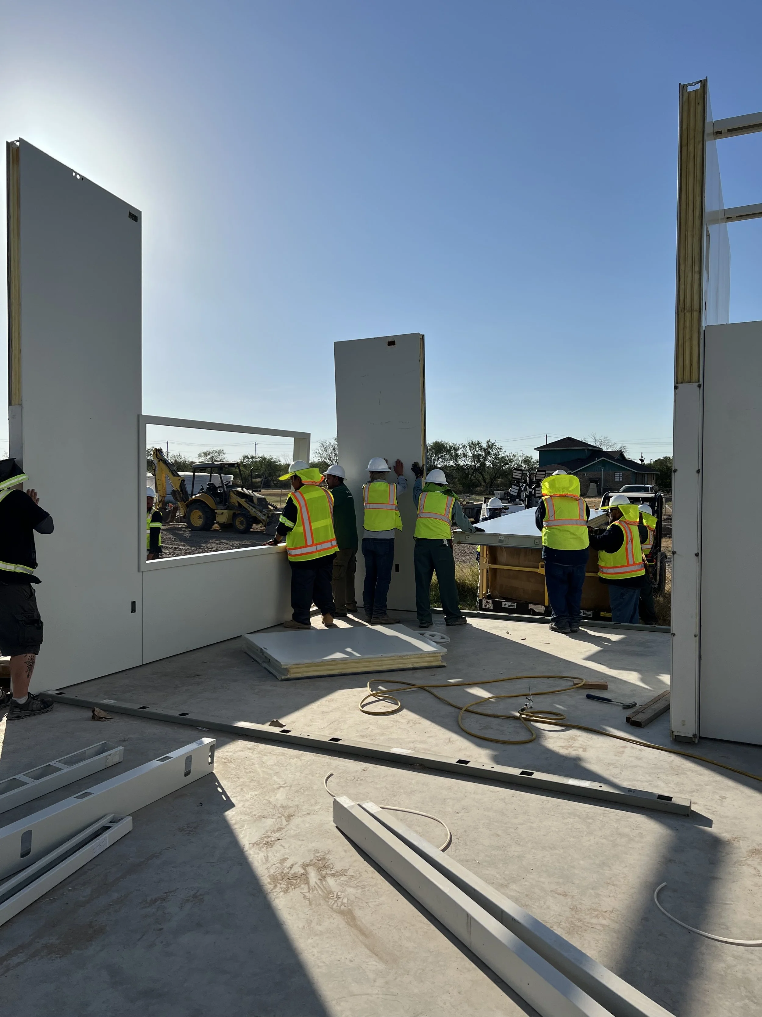 Construction workers in safety vests and helmets assembling large prefabricated wall panels on a building site under a clear sky.